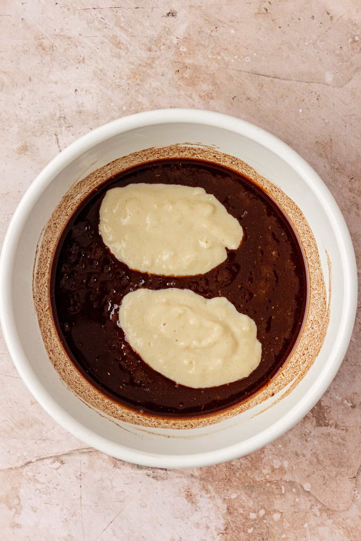 A white bowl containing chocolate batter with two oval-shaped dollops of light-colored batter on top, set on a beige, textured surface.