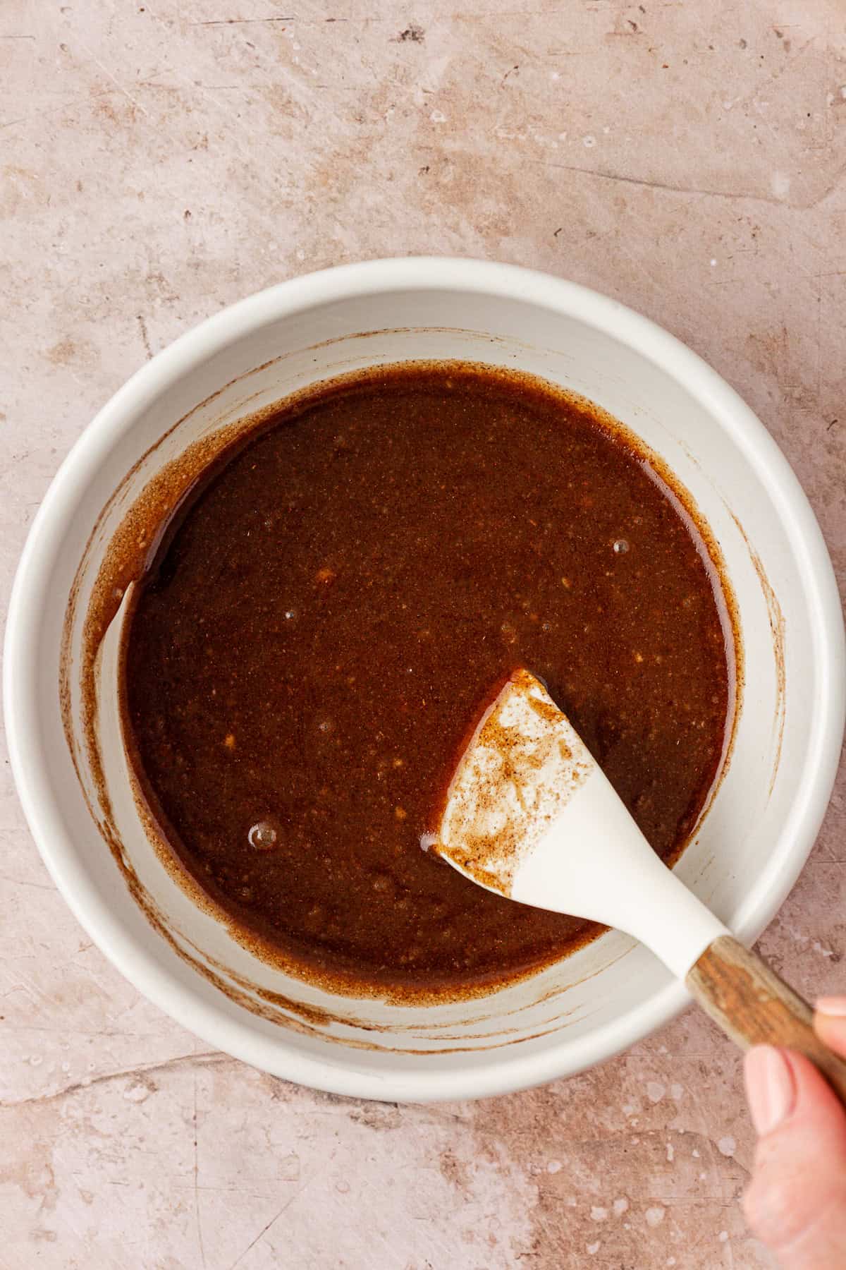 A hand holds a white spatula stirring a thick, dark brown mixture in a white bowl on a light, textured surface.