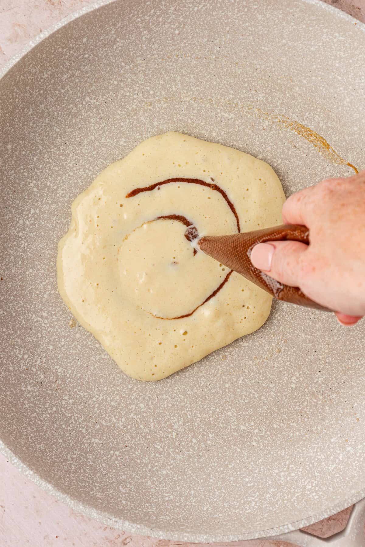A hand pipes a spiral of brown cinnamon filling onto a pancake cooking in a nonstick skillet.