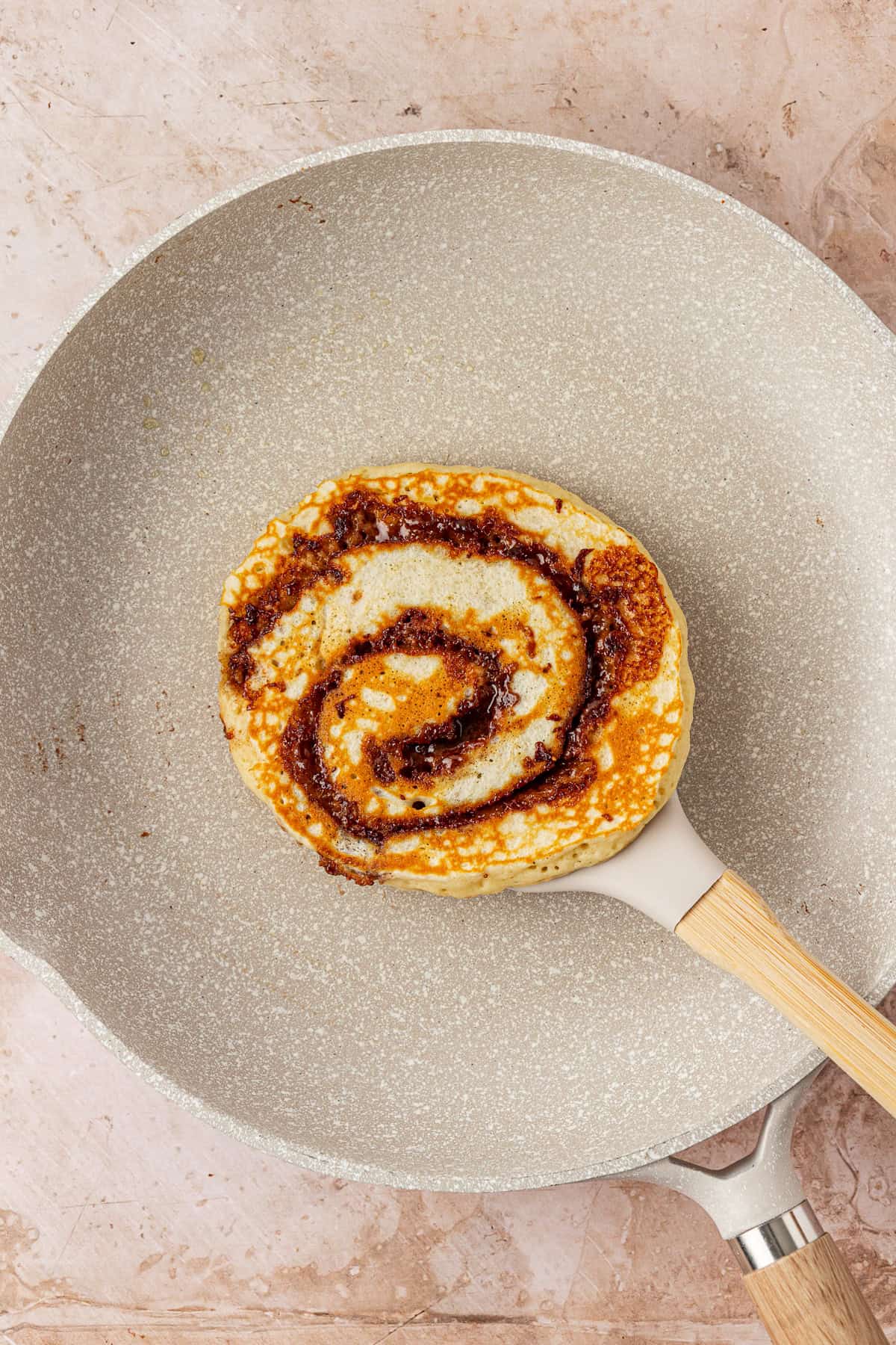 A single pancake with a dark brown swirl pattern cooks in a light-colored nonstick frying pan on a beige countertop.