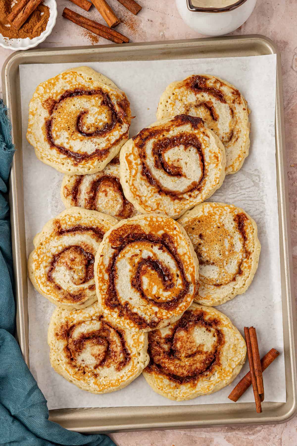 A baking tray lined with parchment paper holds eight cinnamon roll pancakes, each swirled with cinnamon sugar. Cinnamon sticks and a blue cloth are nearby on a beige surface.