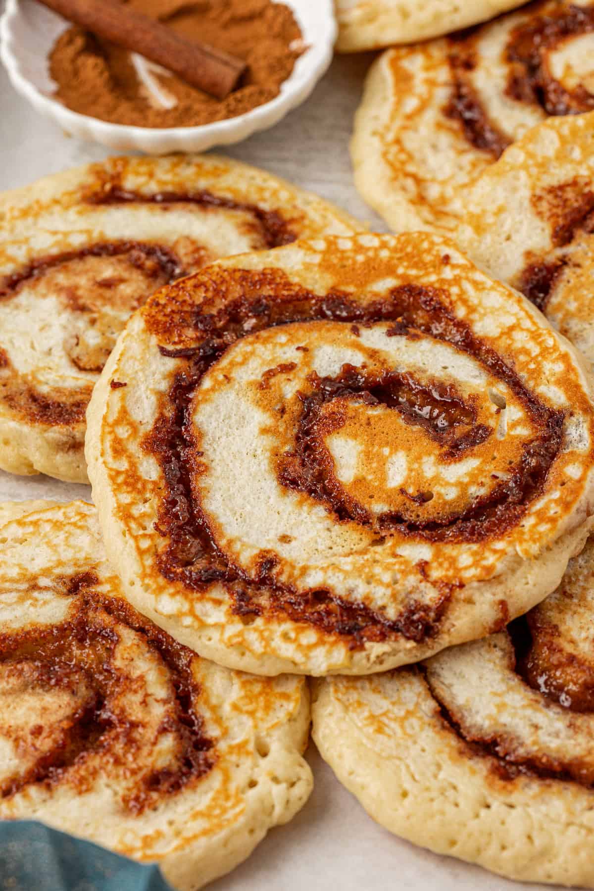 A close-up of several cinnamon swirl pancakes stacked on top of each other, with a small dish of cinnamon sticks and ground cinnamon in the background. The pancakes have golden-brown edges and visible cinnamon filling.