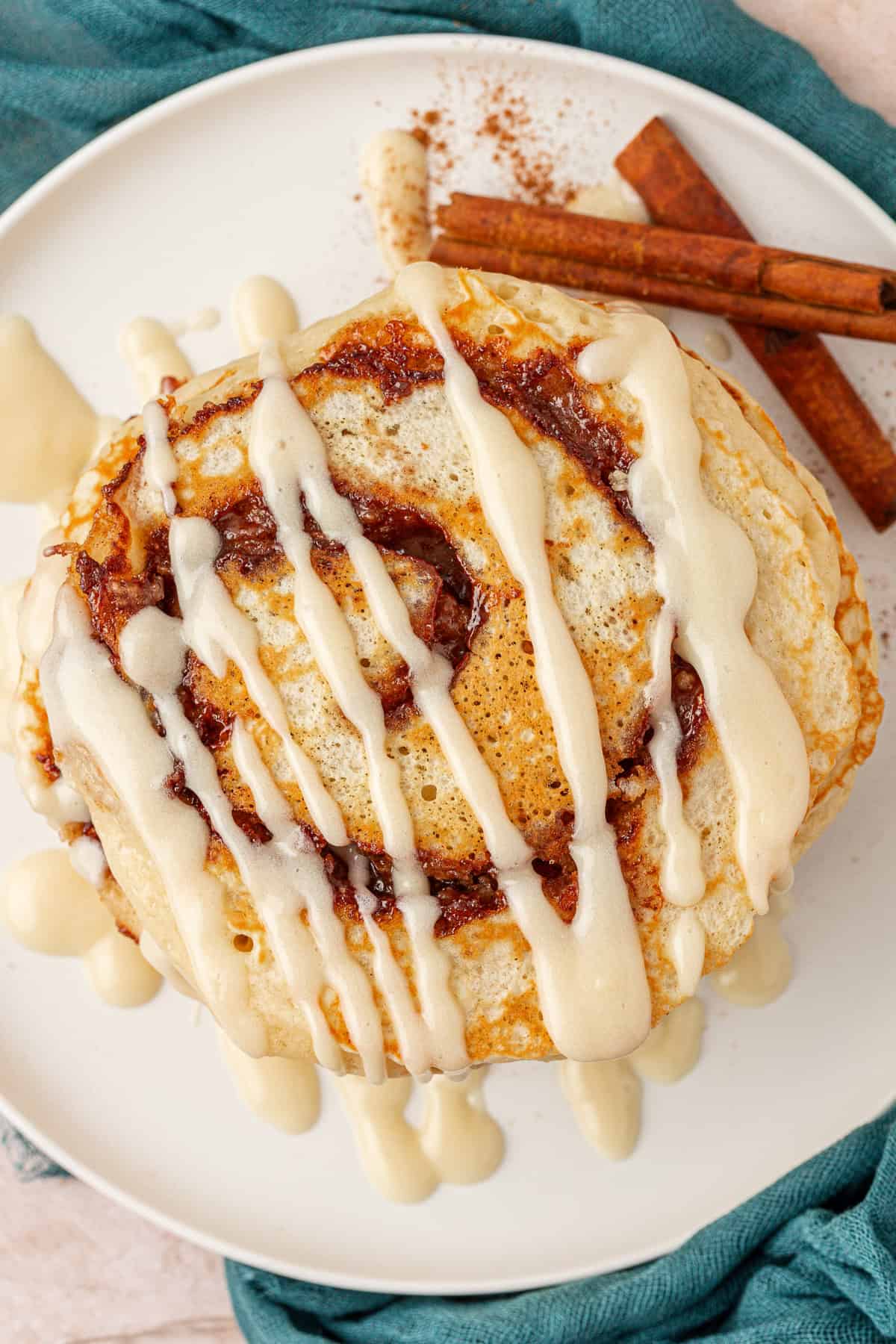 A stack of cinnamon swirl pancakes drizzled with creamy icing sits on a white plate, with two cinnamon sticks and a sprinkle of cinnamon beside them. A teal cloth is visible underneath the plate.