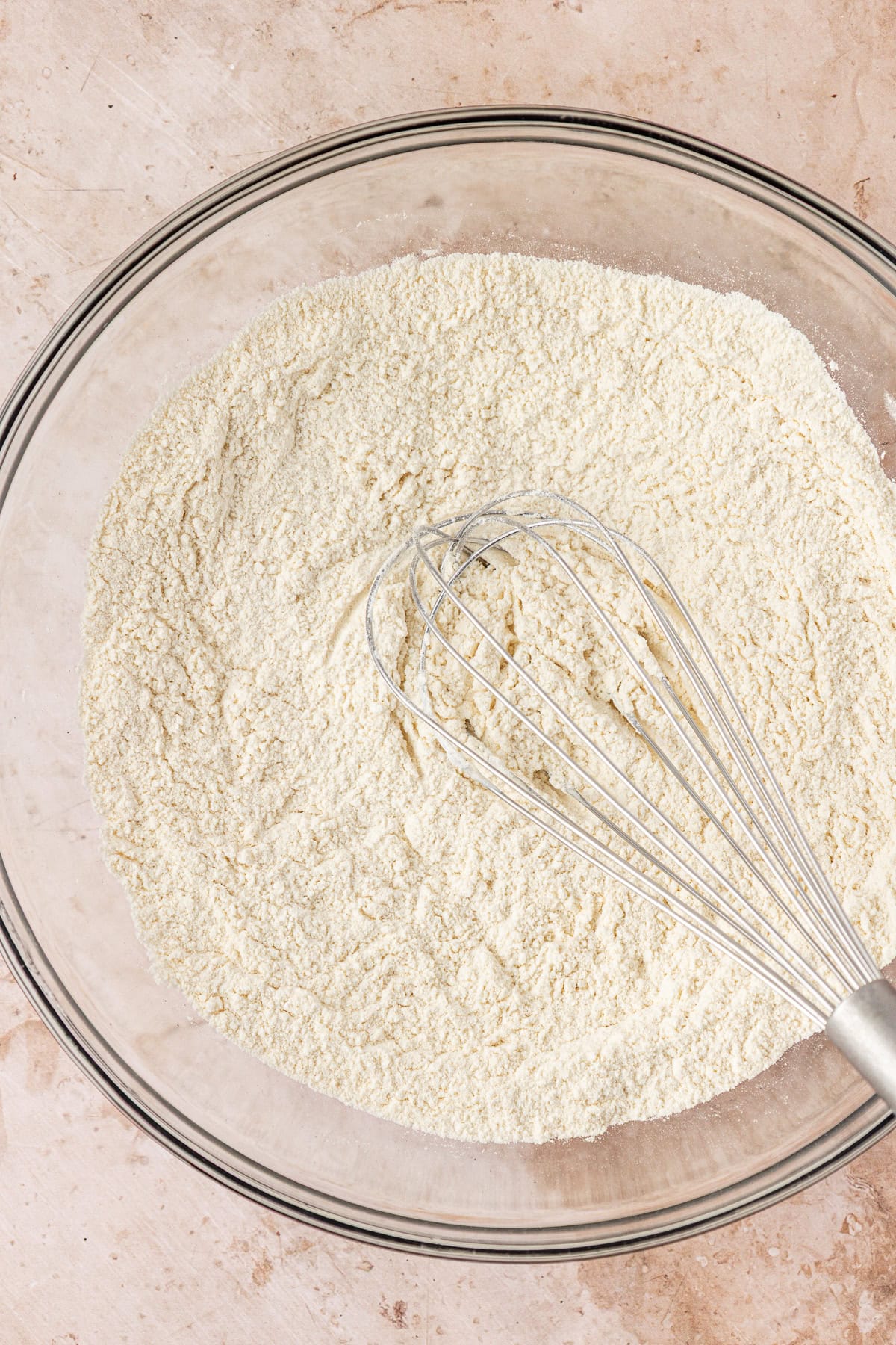 A clear glass bowl filled with flour and a metal whisk resting inside, placed on a light beige countertop.