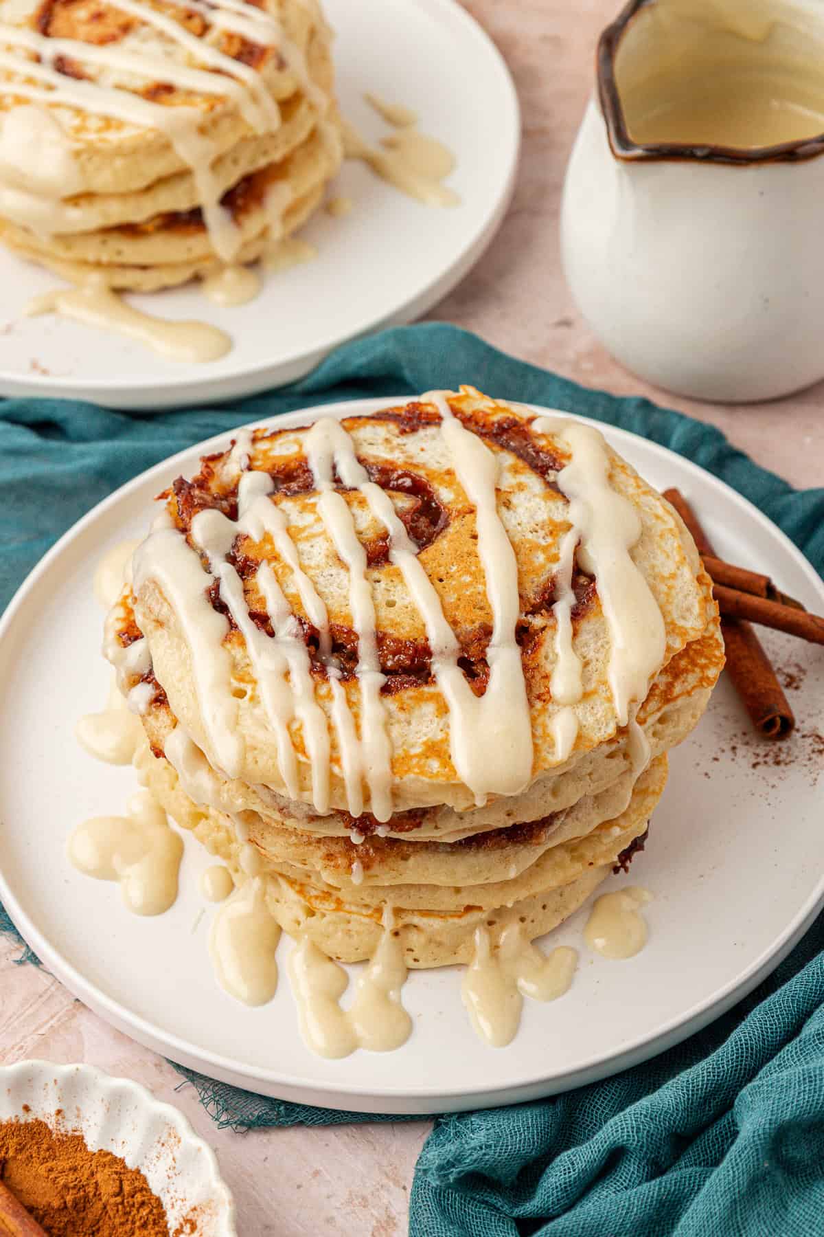 A stack of cinnamon roll pancakes drizzled with icing sits on a white plate. A white pitcher of extra icing and a second plate of pancakes are nearby, with cinnamon sticks and a teal cloth in the background.