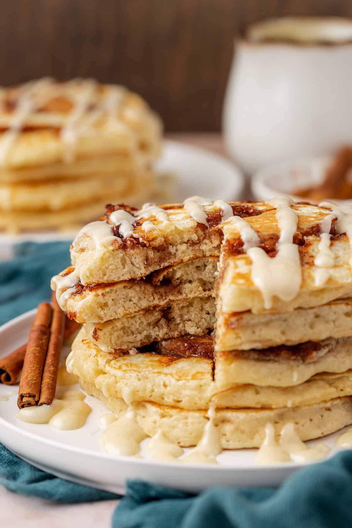 A stack of fluffy pancakes with a cinnamon swirl, drizzled with icing, served on a white plate with cinnamon sticks and a blue napkin. Several pancakes have been cut and are slightly separated to show the soft interior.