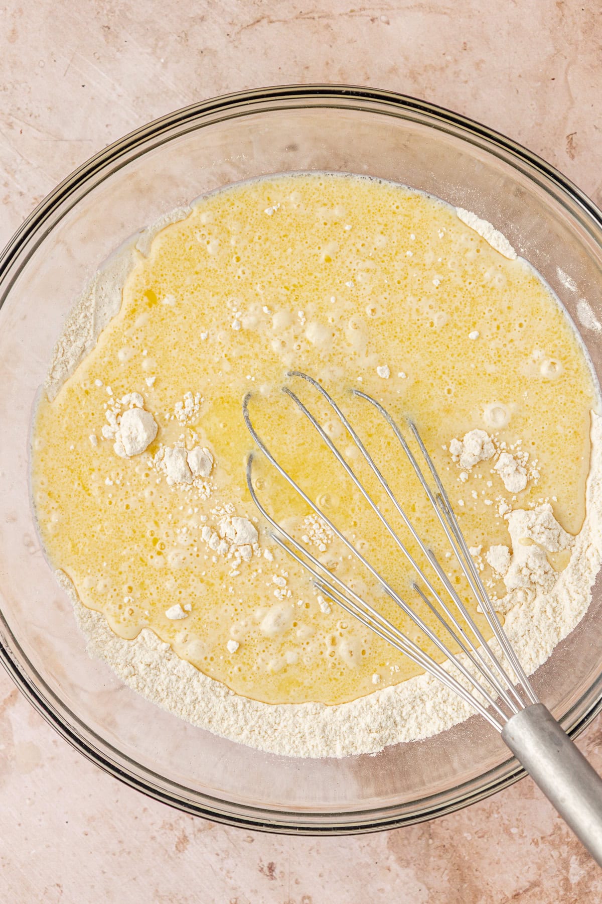 A glass bowl on a countertop contains flour and a yellow liquid mixture, partially combined. A metal whisk rests inside the bowl, ready for mixing.
