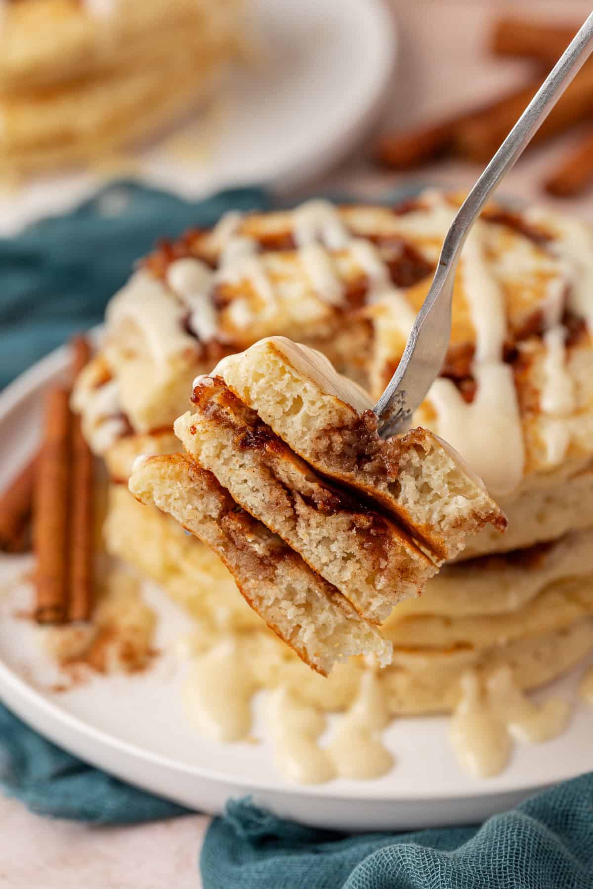 A fork holds up a bite of fluffy cinnamon swirl pancakes drizzled with icing, with a stack of pancakes and cinnamon sticks on a plate in the background.
