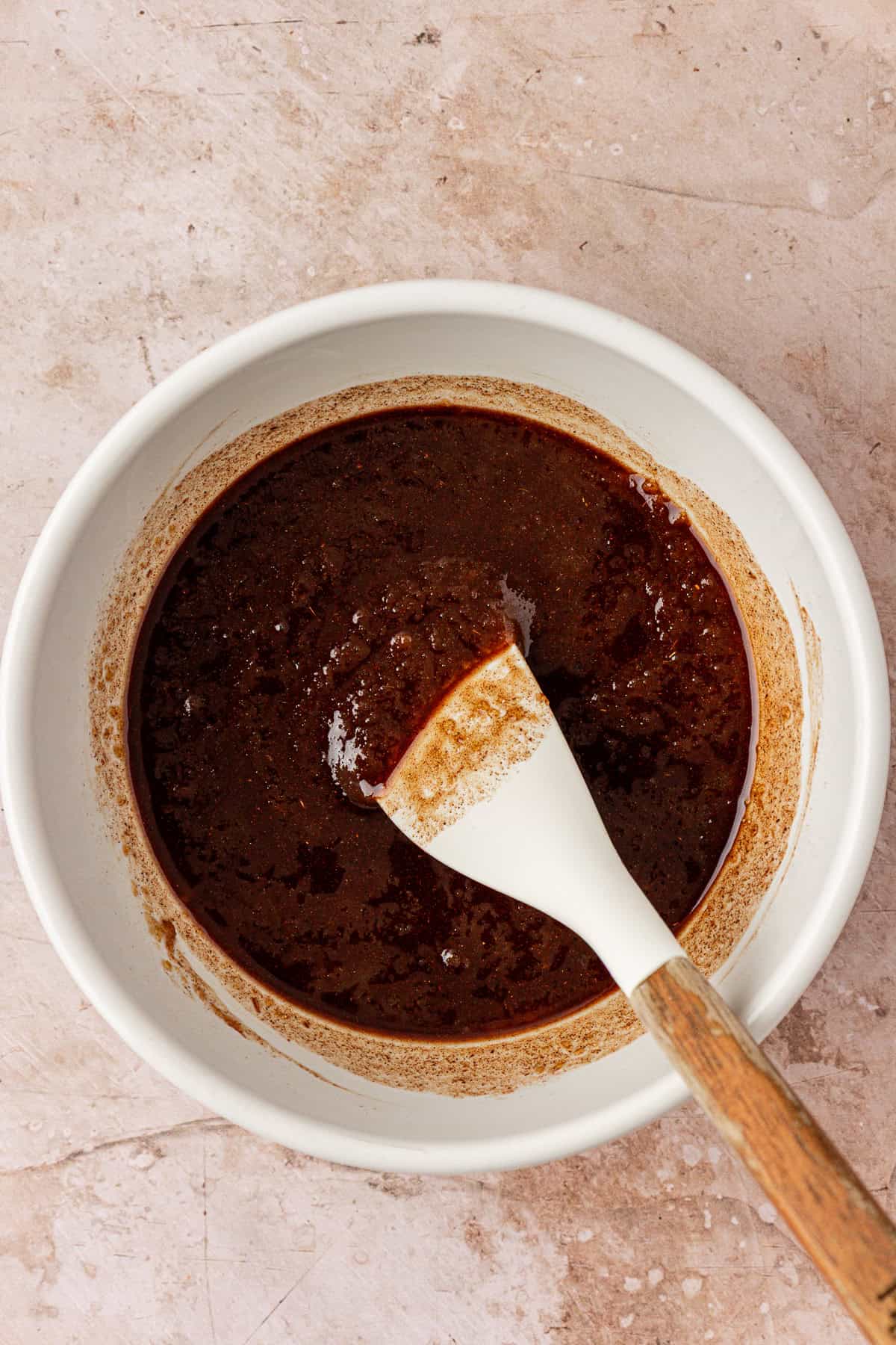 A white bowl containing a thick, dark brown mixture is being stirred with a white and wooden spatula on a light brown textured surface.