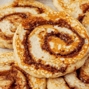 Close-up of several round, swirled cinnamon rolls with a golden-brown baked crust and visible cinnamon filling, overlapping each other on a light background.