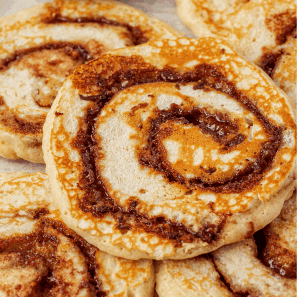 Close-up of several round, swirled cinnamon rolls with a golden-brown baked crust and visible cinnamon filling, overlapping each other on a light background.