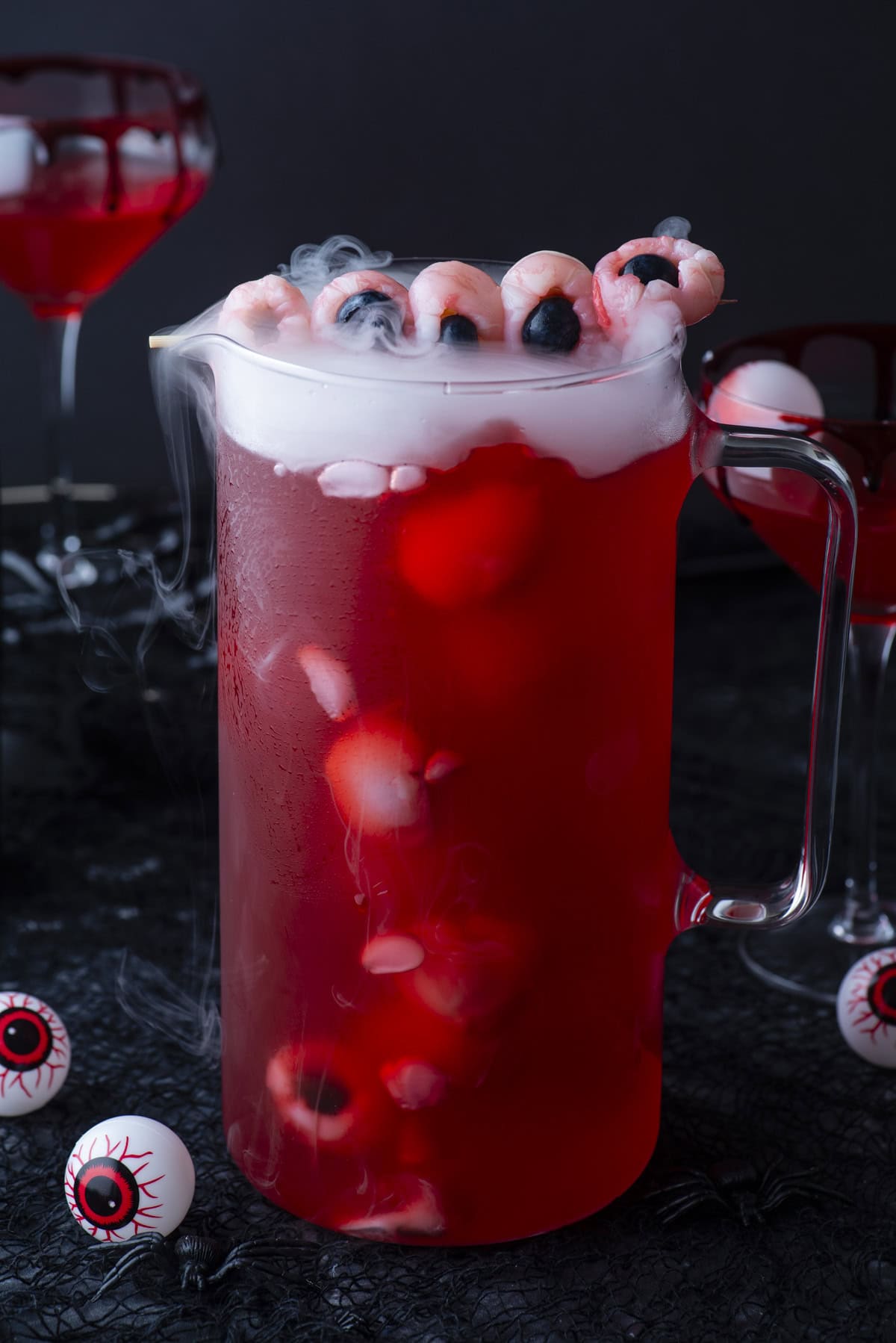 A glass pitcher of red punch with floating lychee eyeballs, blueberries, and dry ice, surrounded by plastic eyeballs and cocktail glasses, creating a spooky Halloween-themed drink scene.