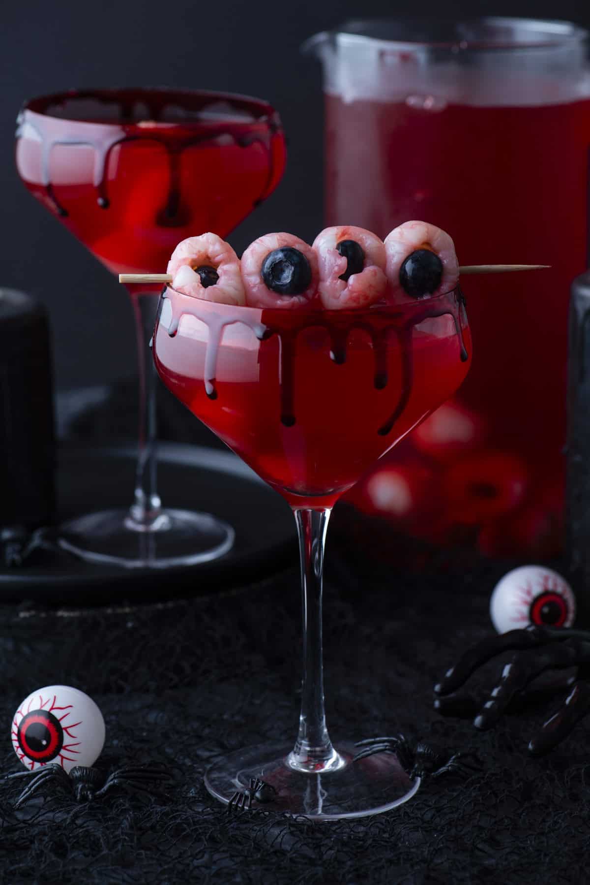 A spooky red cocktail in a glass with black syrup dripping down the rim, garnished with lychees and blueberries to look like eyeballs on a skewer. A pitcher of red drink and Halloween decorations are in the background.