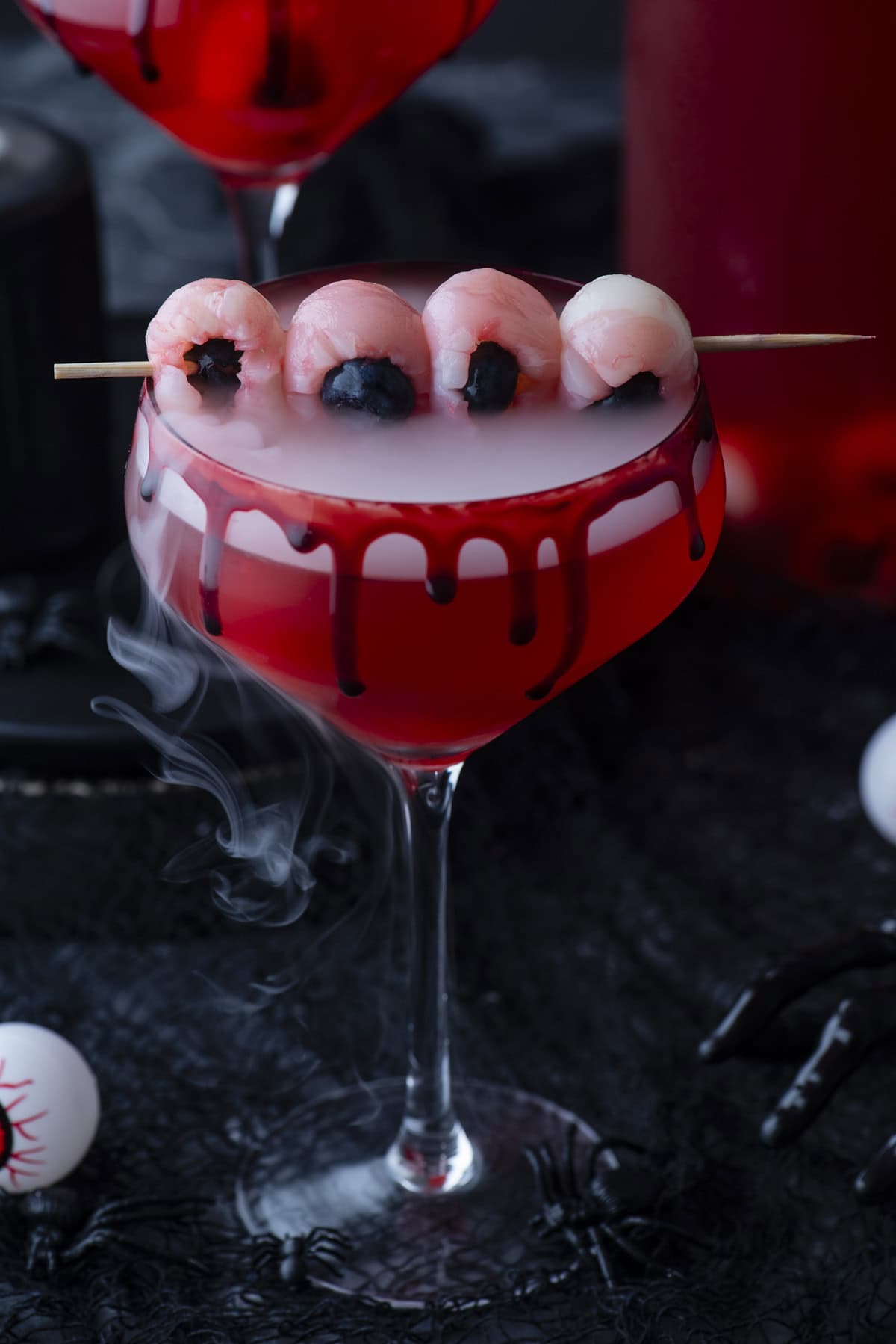 A spooky red cocktail in a glass with black syrup dripping down the rim, topped with lychee “eyeballs” on a skewer and wisps of fog, set on a dark background with Halloween decorations.