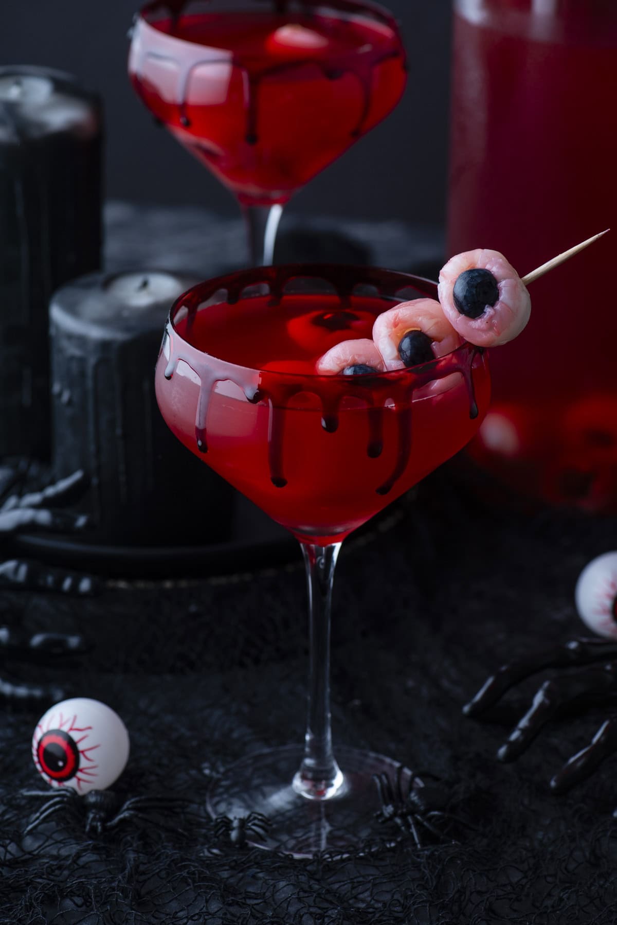 A spooky red cocktail in a glass with black drip detail, garnished with eyeball-shaped treats on a skewer. Black candles, a plastic hand, and fake eyeballs add to the Halloween theme.