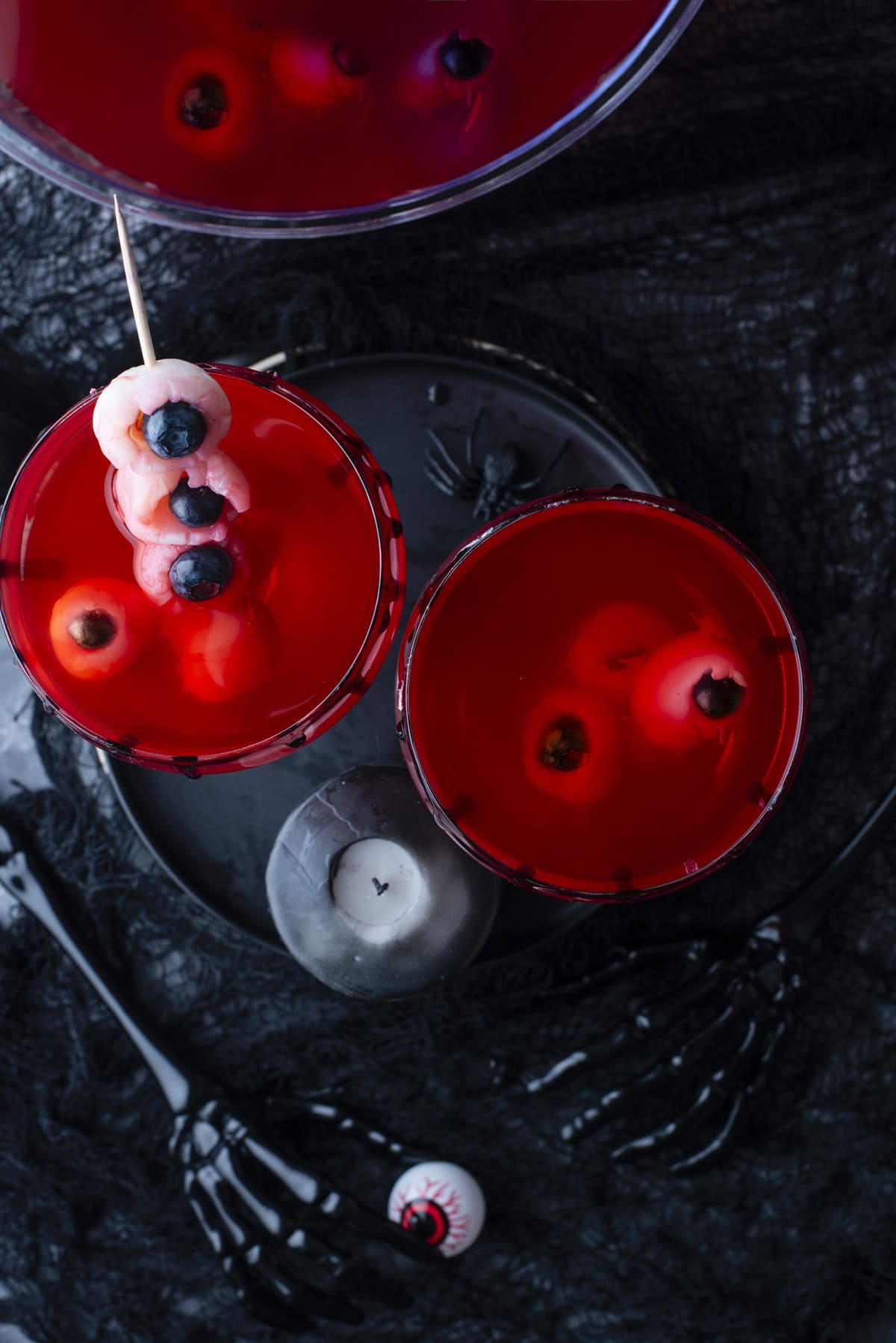 Overhead view of red drinks in glasses with eyeball-shaped garnishes, surrounded by black Halloween-themed decorations and skeleton hand utensils. A black candle and a fake eyeball are also visible.