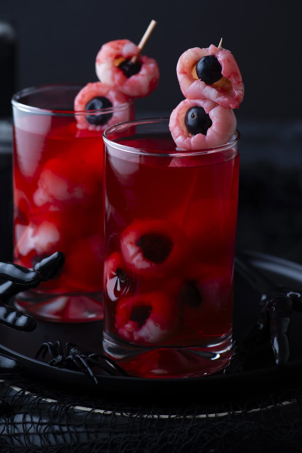 Two glasses filled with red liquid, each containing peeled lychees stuffed with blueberries to resemble eyeballs, garnished with skewered lychee eyeballs on top. A black plastic hand decorates the tray beneath.