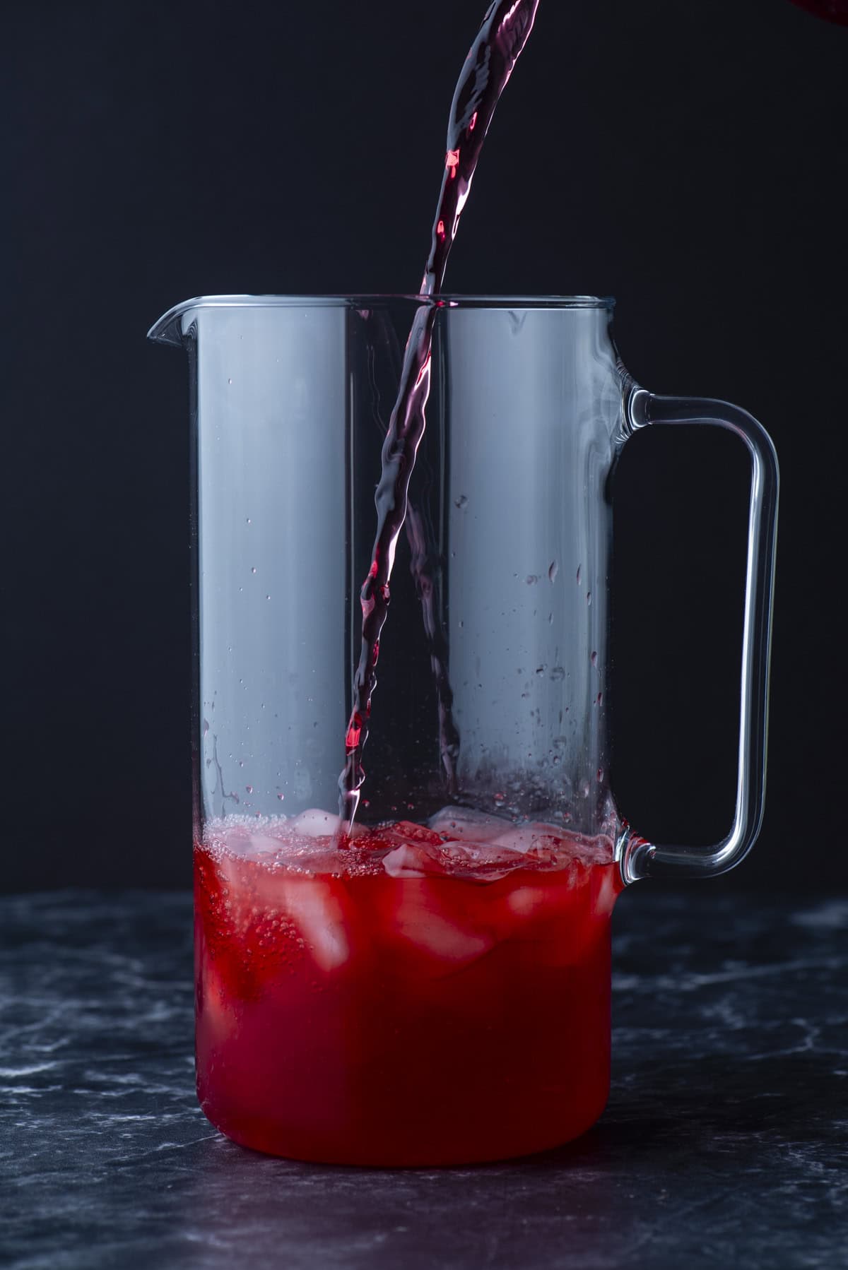 A clear glass pitcher with ice is being filled with a red liquid, possibly juice or punch, against a dark background.