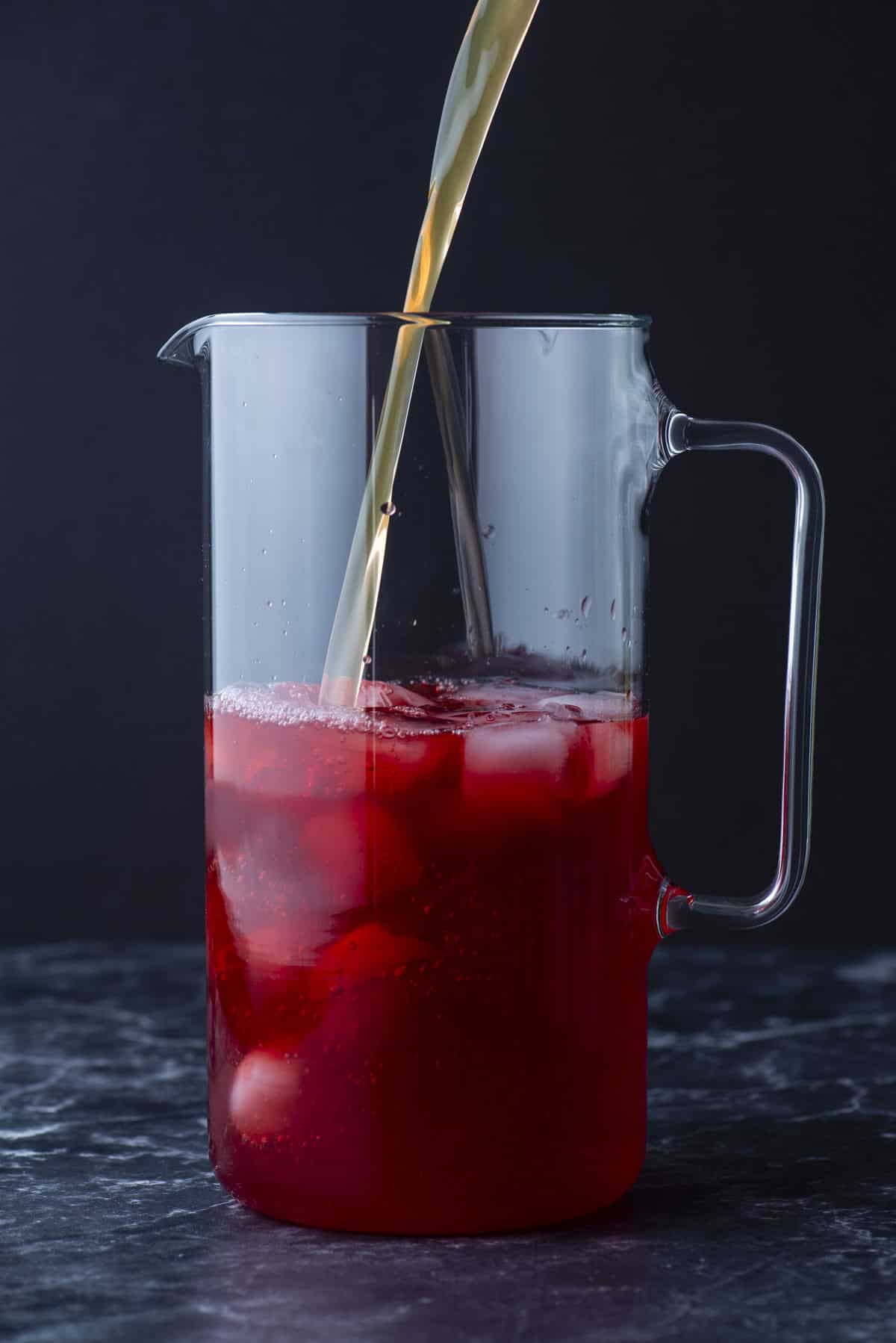 A clear glass pitcher filled with ice and red liquid, with a stream of light-colored liquid being poured in, against a dark background.