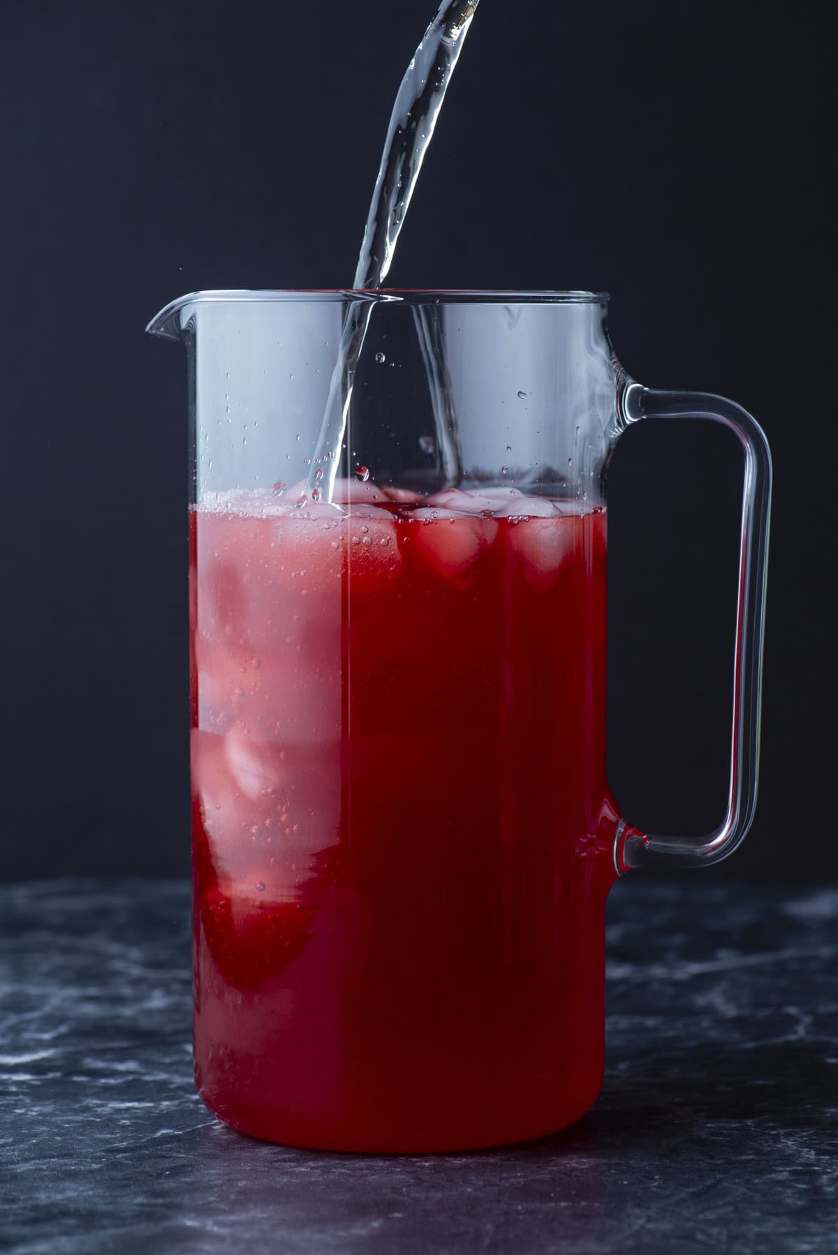A clear glass pitcher filled with red liquid and ice is being topped up with water. The pitcher is set on a dark, textured surface with a black background.