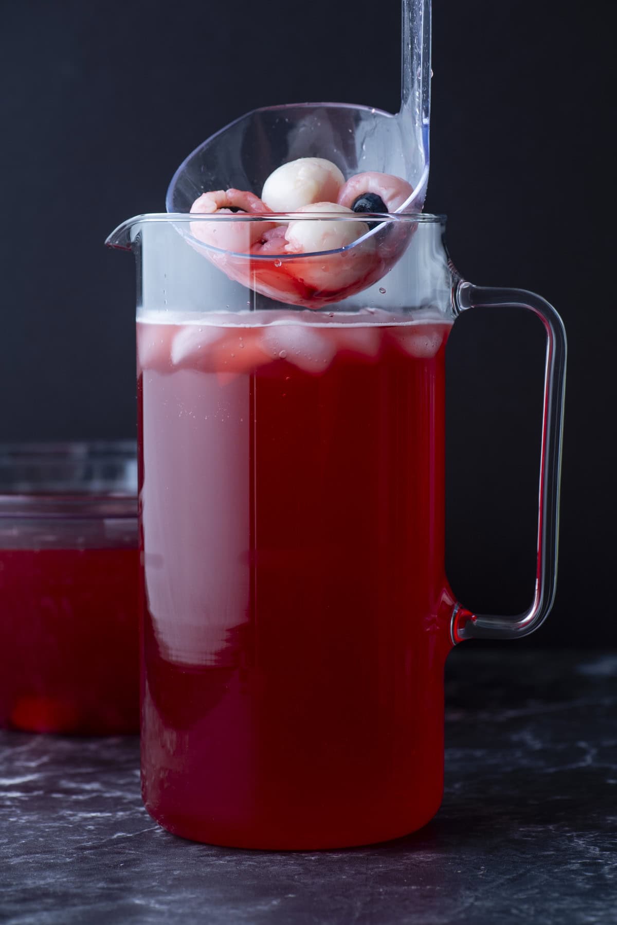 A glass pitcher filled with red punch and ice, topped with peeled lychees and blueberries. A clear ladle holds more lychees and blueberries above the pitcher. The background is dark and minimal.