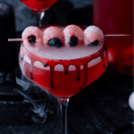 A red cocktail with a smoky effect is served in a glass with black and red syrup dripping down the rim. Four lychee and blueberry eyeballs are skewered on top, creating a spooky Halloween drink.