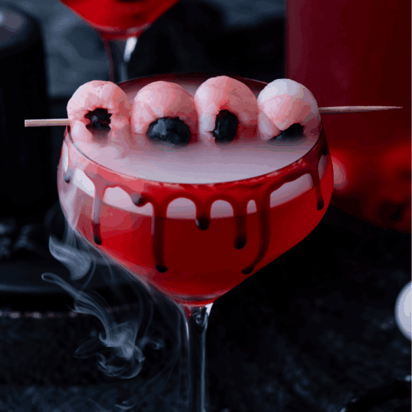 A red cocktail with a smoky effect is served in a glass with black and red syrup dripping down the rim. Four lychee and blueberry eyeballs are skewered on top, creating a spooky Halloween drink.
