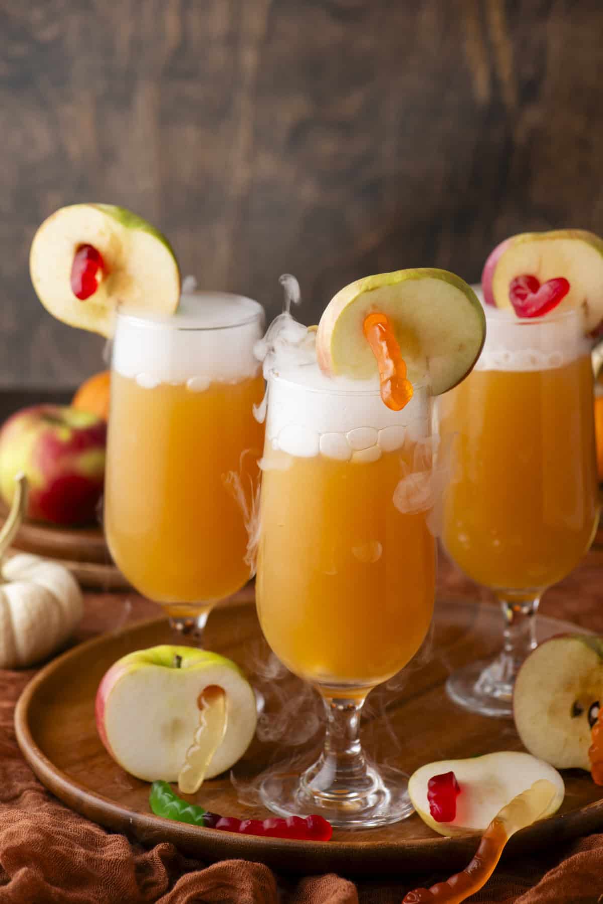 Three orange cocktails in tall glasses, garnished with apple slices and gummy worms, emit wisps of smoke. The drinks sit on a wooden tray with more apples and gummy worms nearby, creating a festive autumn setting.
