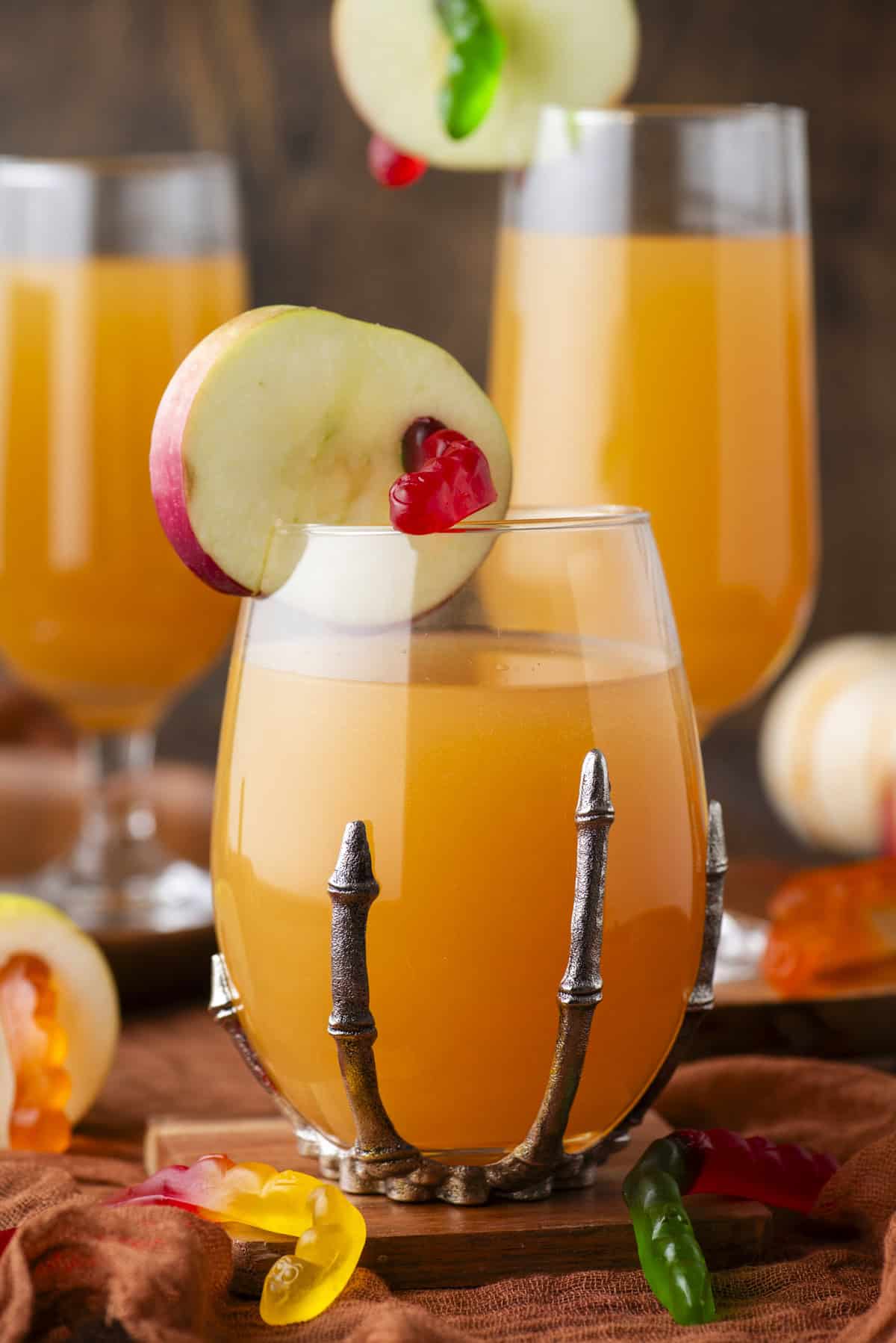 A glass of orange juice is garnished with a red apple slice and gummy candies. The glass is held in a decorative metal holder shaped like a skeletal hand. Two more similar drinks and gummies are visible in the background.
