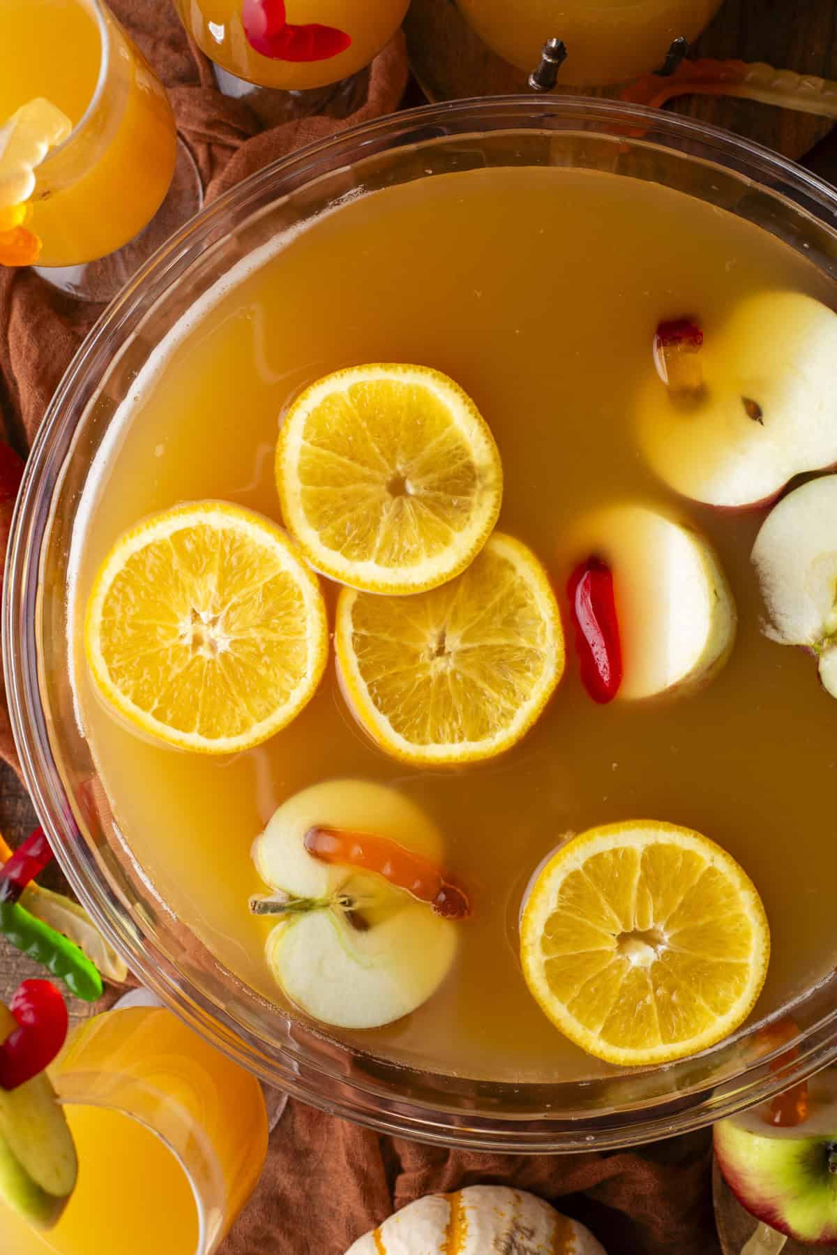 A bowl of orange-colored punch topped with sliced oranges and apples, with glasses of the drink and autumn decor around it.