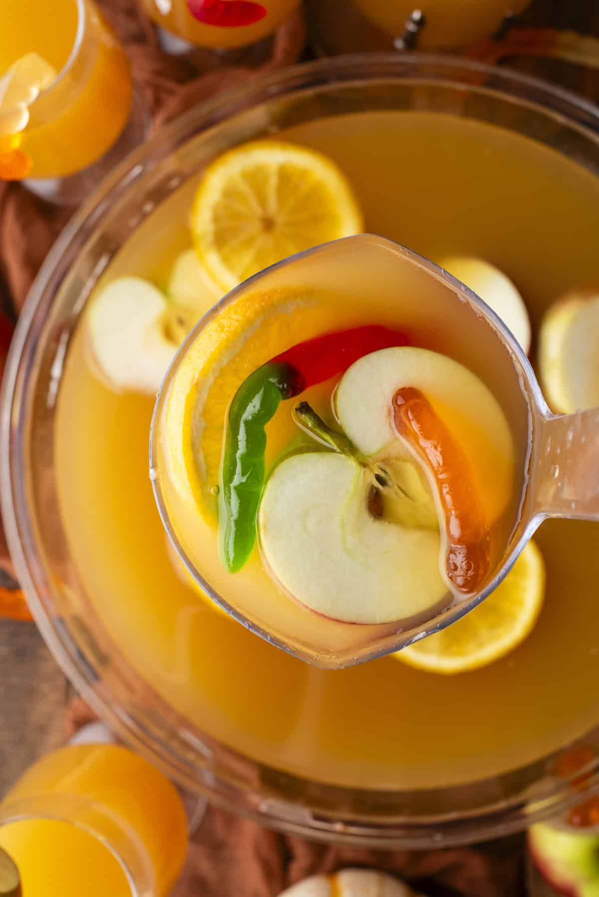 A ladle scooping fruit punch from a bowl, containing apple slices, orange slices, and whole green and red chili peppers. The punch is orange and surrounded by glasses filled with the same drink.