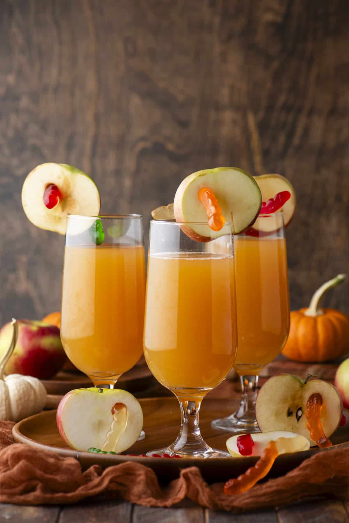 Three glasses of orange-colored drinks are garnished with apple slices and gummy worms. The glasses are on a wooden tray, surrounded by apples, a small pumpkin, and rustic fabric, set against a wooden background.