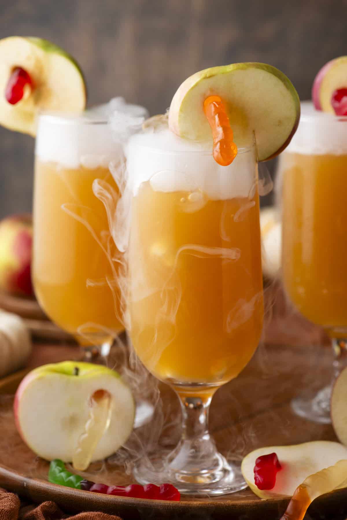 Three glasses of orange punch with foamy tops, garnished with apple slices and gummy worms, surrounded by apple slices and vapor, creating a festive and playful atmosphere.