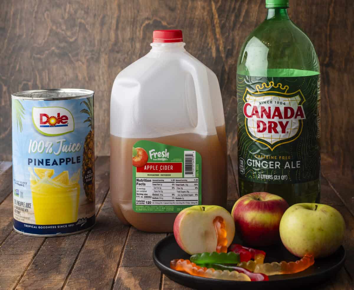 A can of Dole pineapple juice, a jug of apple cider, a bottle of Canada Dry ginger ale, and a plate with apples and gummy worms are displayed on a wooden surface against a wood-textured background.