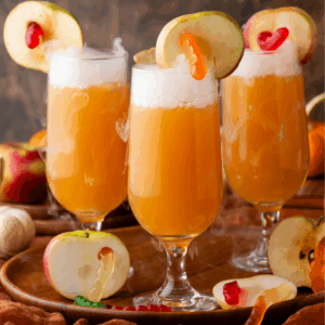 Three glasses of orange punch topped with foam, each garnished with a slice of apple and a gummy worm. Whole and sliced apples, plus more gummy worms, are arranged on a wooden tray with a warm, rustic background.