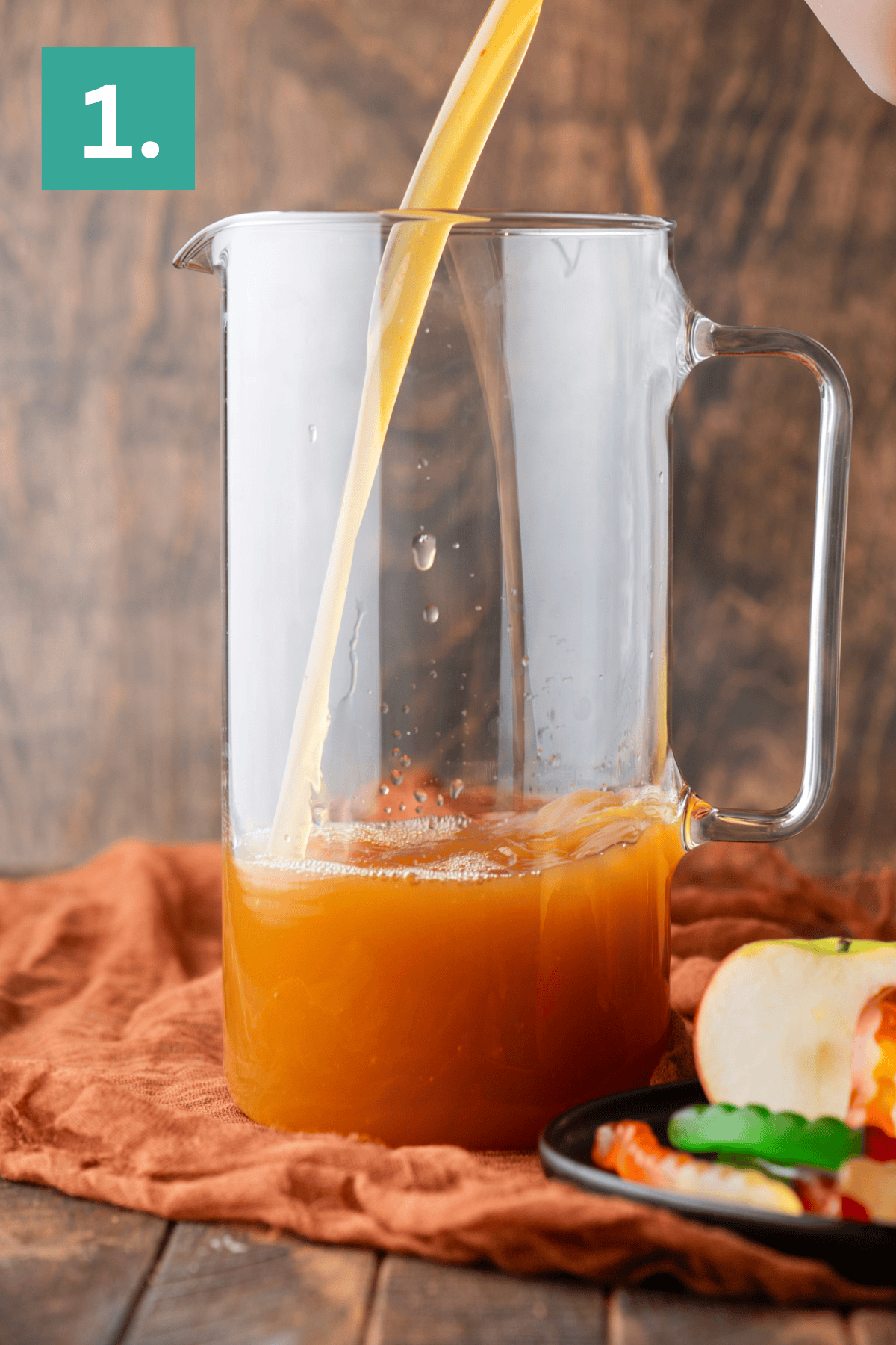 A glass pitcher is being filled with orange-colored juice. The pitcher sits on an orange cloth, with sliced apples and gummy worms on a plate nearby. A green box with the number 1 appears in the top left corner.