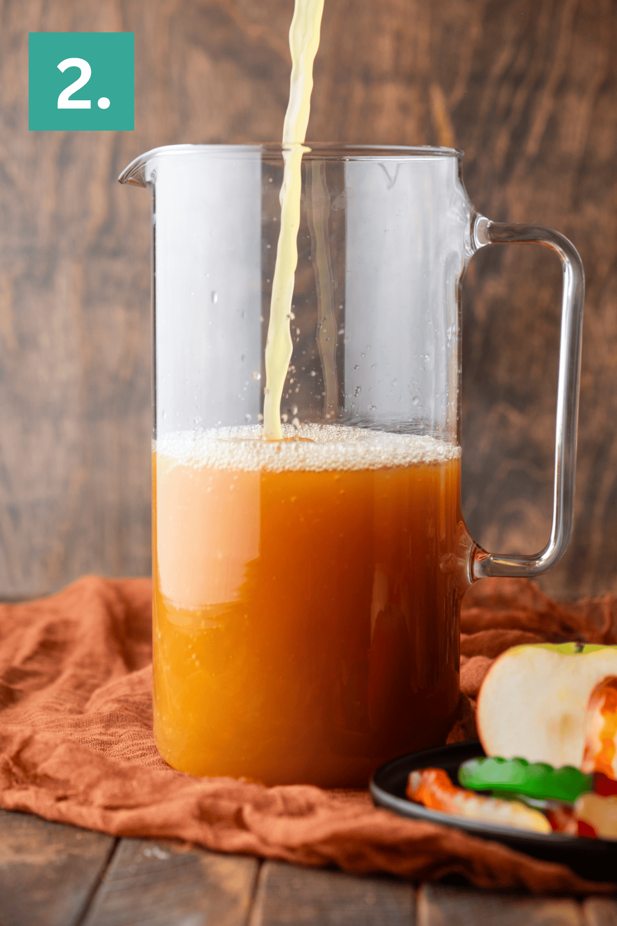 A glass pitcher is being filled with orange-brown juice. The pitcher sits on a brown cloth with an apple and gummy worms on a small plate nearby. A green box with the number 2 is in the upper left corner.