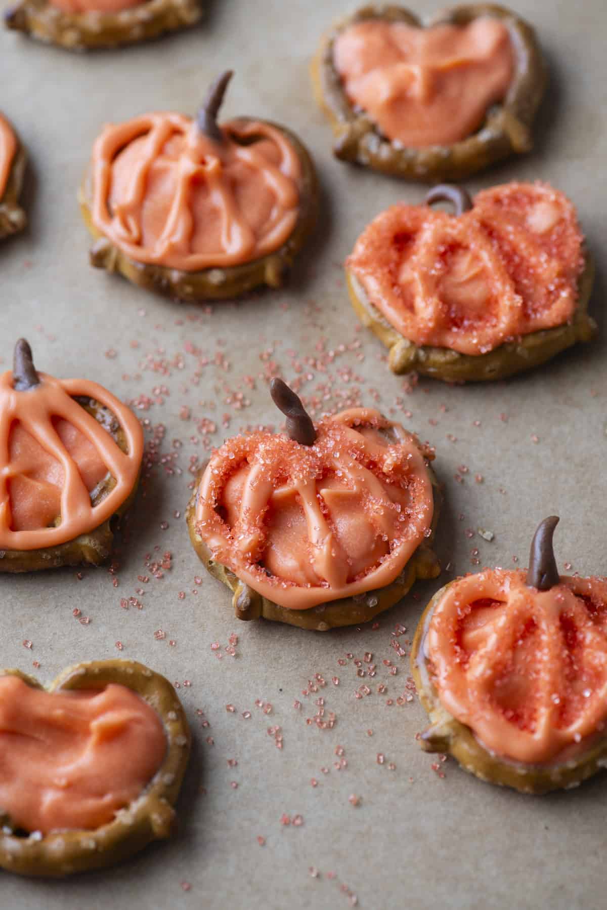 Pumpkin-shaped pretzel treats topped with orange icing and sprinkles are arranged on parchment paper. Some feature chocolate stems and decorative icing outlining pumpkin details.