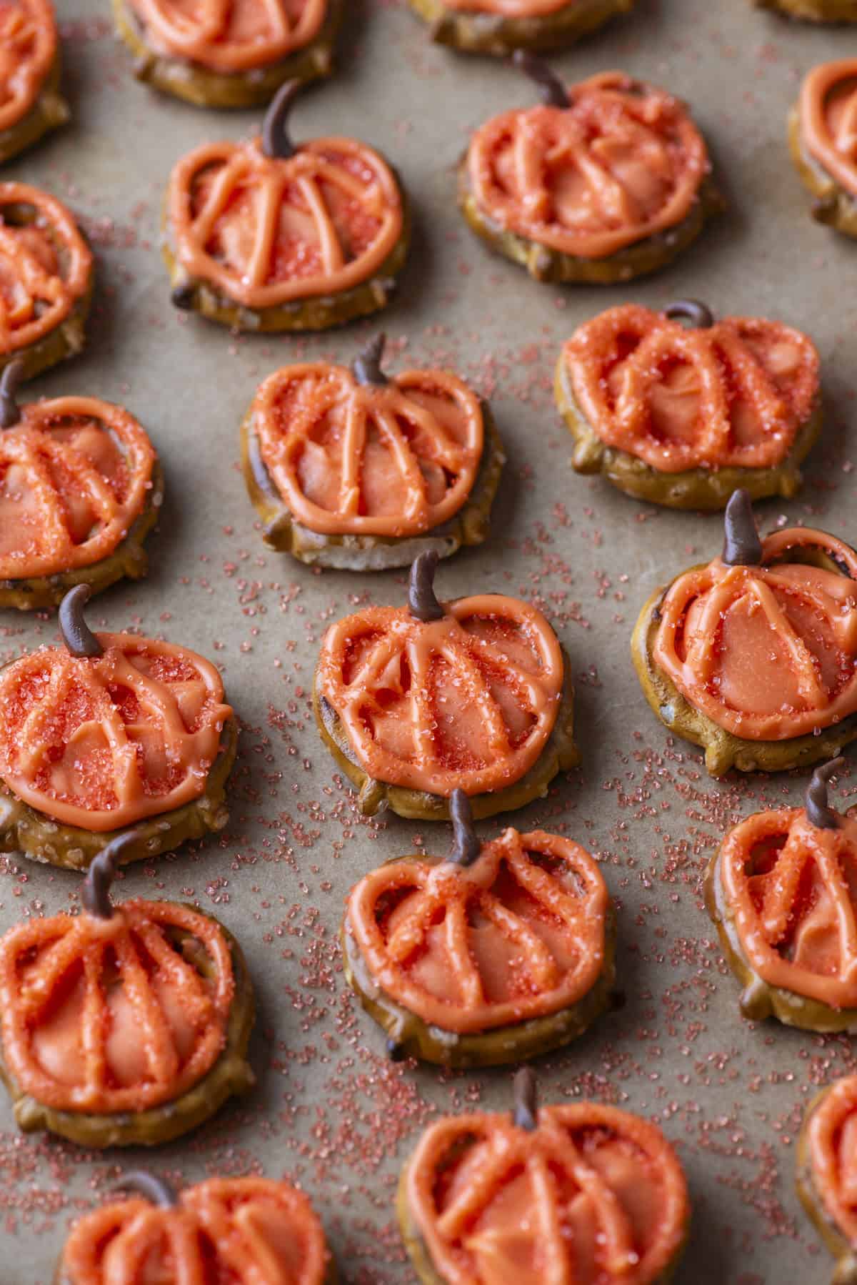 Pumpkin-shaped cookies with orange icing and red sprinkles are arranged on a baking sheet. Each cookie has a small brown stem, and red sugar crystals are scattered around them.