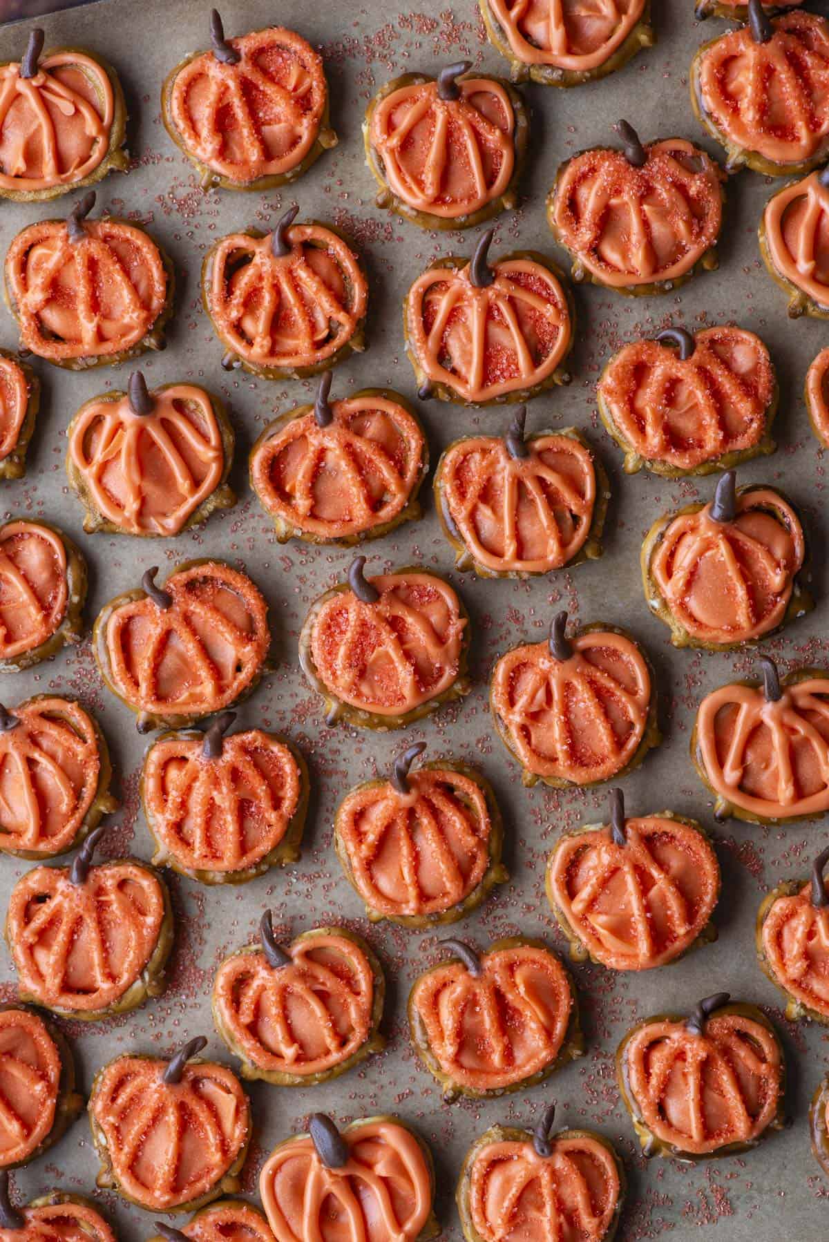 Rows of pumpkin-shaped cookies decorated with orange icing and sprinkles, each with a small chocolate stem, arranged closely together on a baking sheet.
