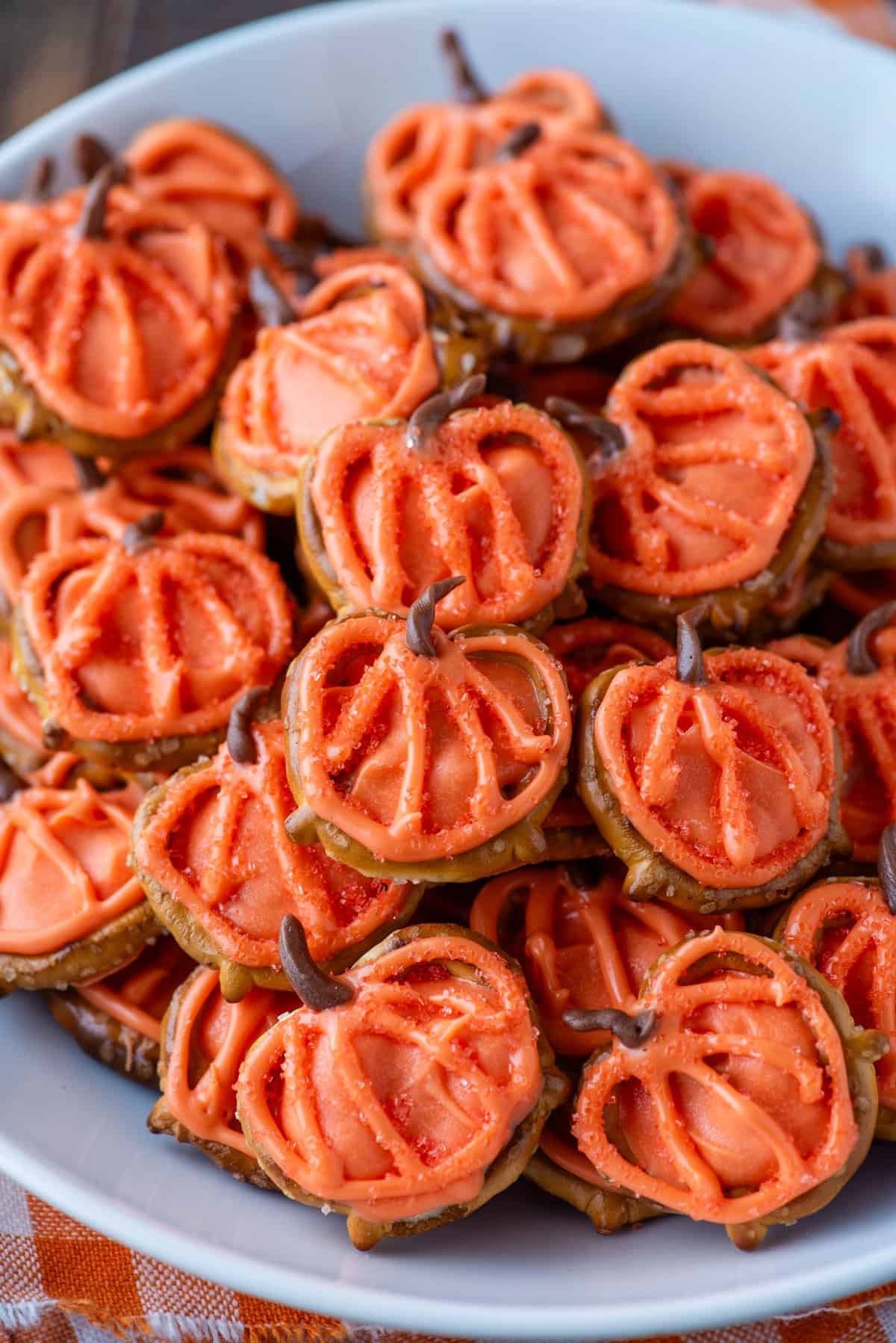A plate filled with pumpkin-shaped treats made from orange icing on pretzels, decorated with chocolate stems and orange sugar sprinkles, arranged in a white bowl.