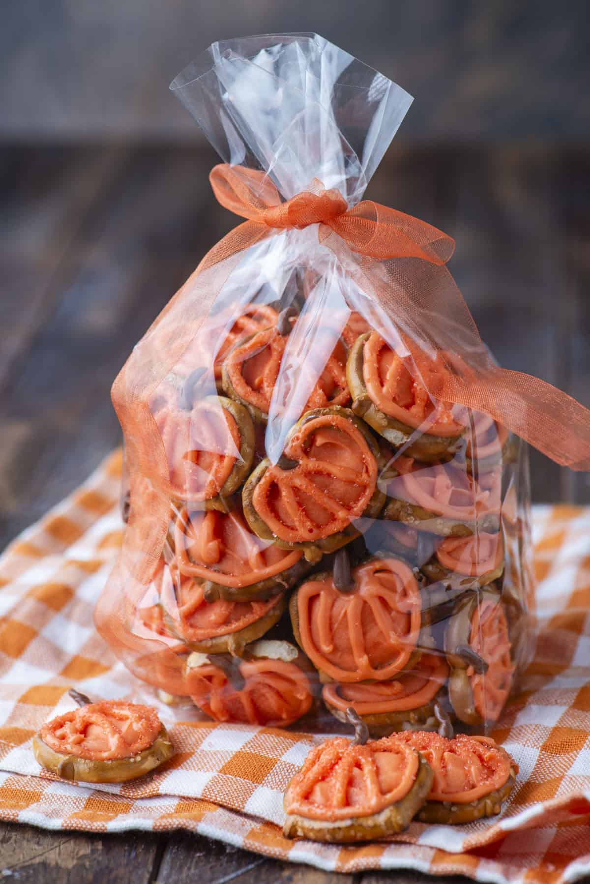 A clear plastic bag tied with an orange ribbon is filled with pretzel snacks topped with orange frosting. The bag sits on an orange-and-white checkered cloth, with a few snacks placed outside the bag.