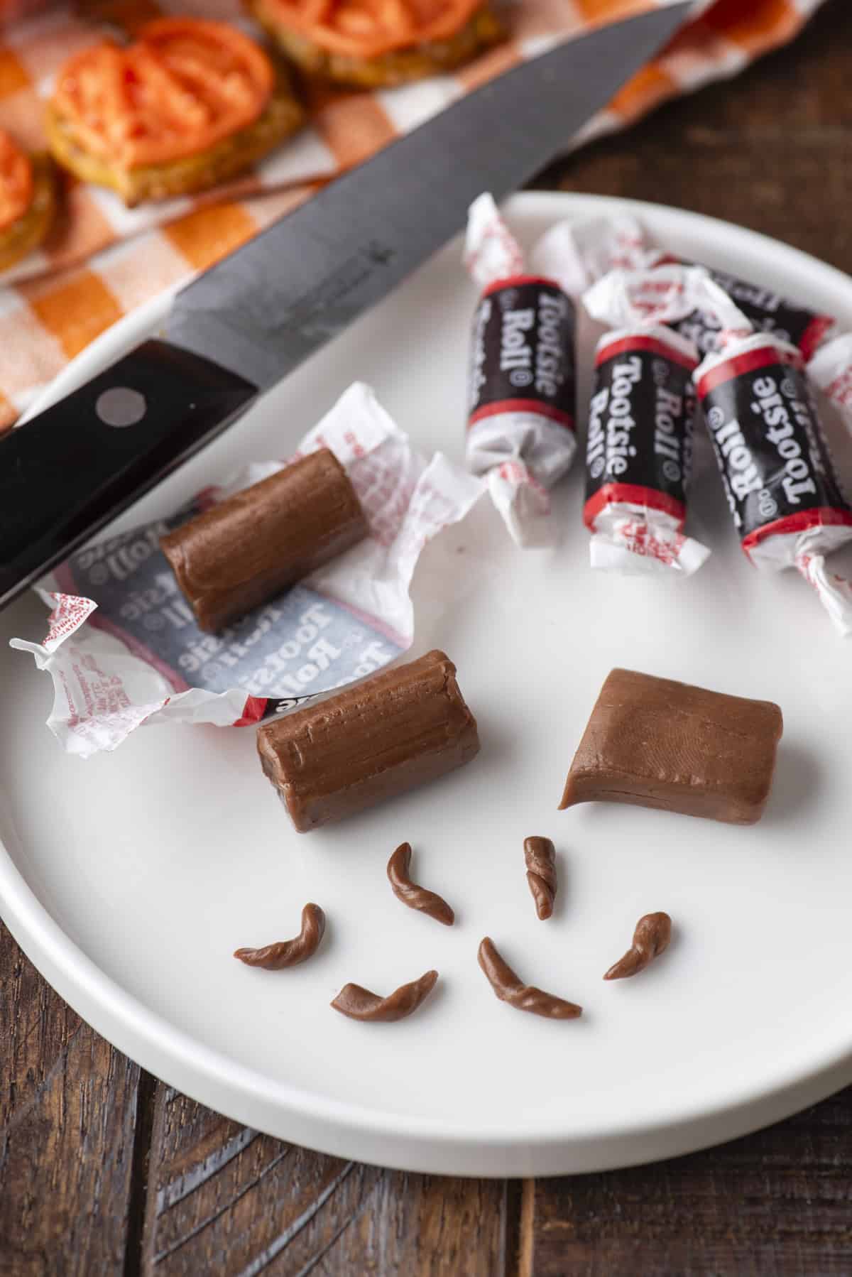 A white plate with unwrapped and wrapped Tootsie Rolls, some cut in pieces, and small pieces shaped like poop. A knife lies nearby on a checkered napkin. Everything is on a wooden table.