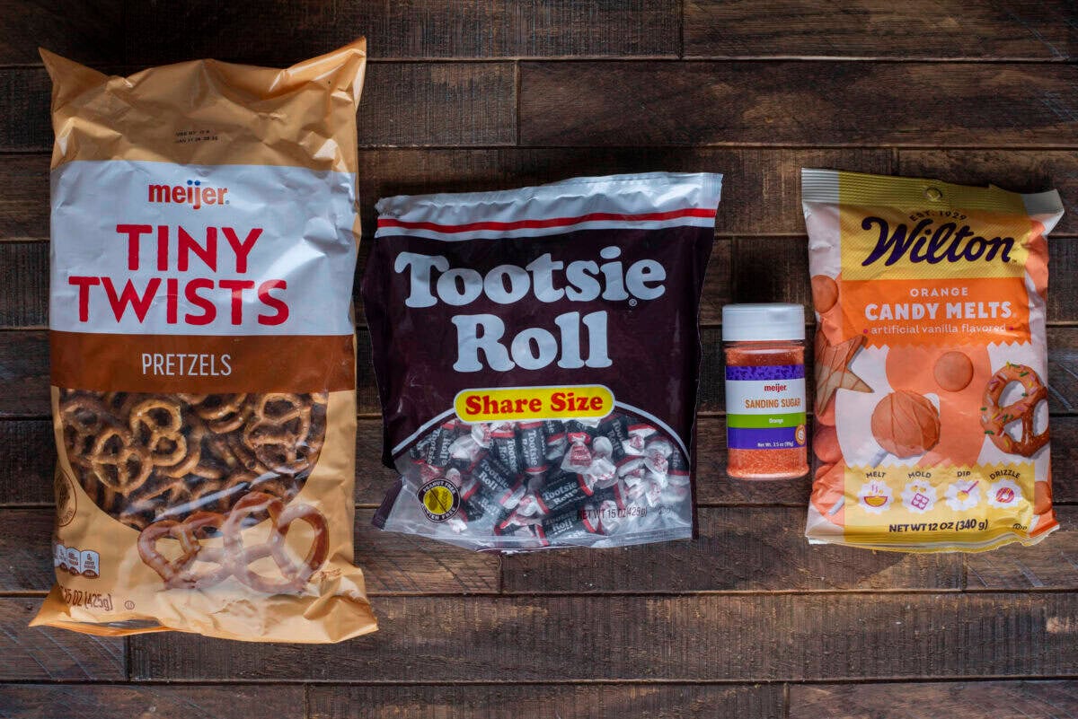 A bag of Meijer Tiny Twists pretzels, a share-size bag of Tootsie Roll candies, a container of orange sanding sugar, and a bag of Wilton orange candy melts on a wooden surface.