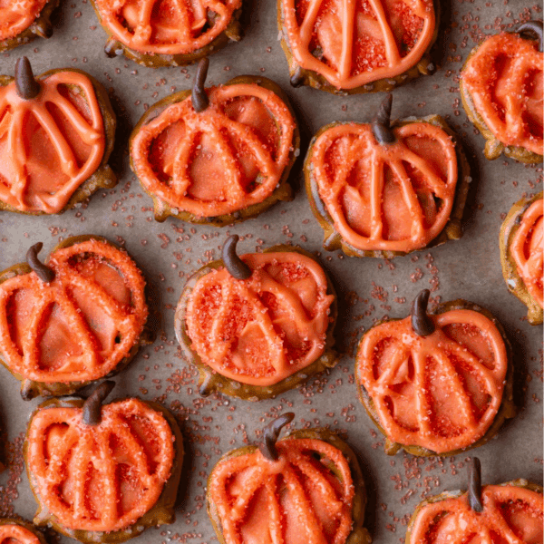 Pumpkin-shaped sugar cookies decorated with orange icing and sprinkles, arranged in rows on parchment paper. Each cookie has a chocolate stem and piped lines imitating a pumpkin’s shape.