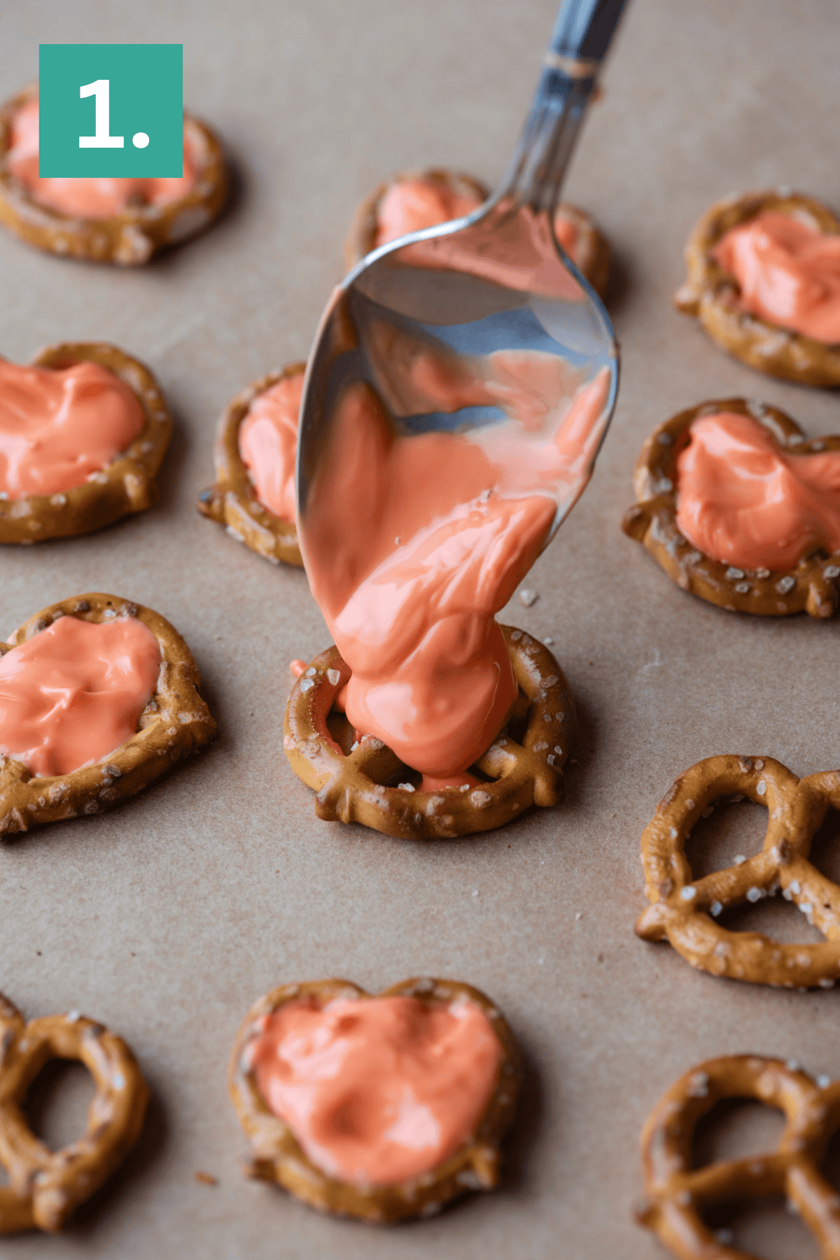 A spoon spreads melted orange-colored candy onto a small pretzel on parchment paper, surrounded by other pretzels already covered in the same candy coating. A green box with “1.” is in the top left corner.