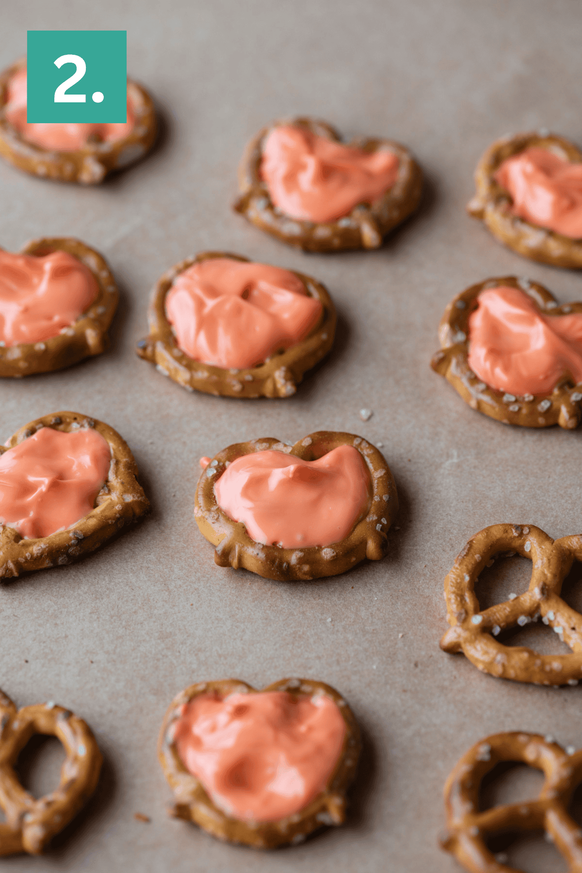 Pretzel twists spread with orange icing are arranged on a brown surface. A green square with a white number 2. appears in the top left corner.