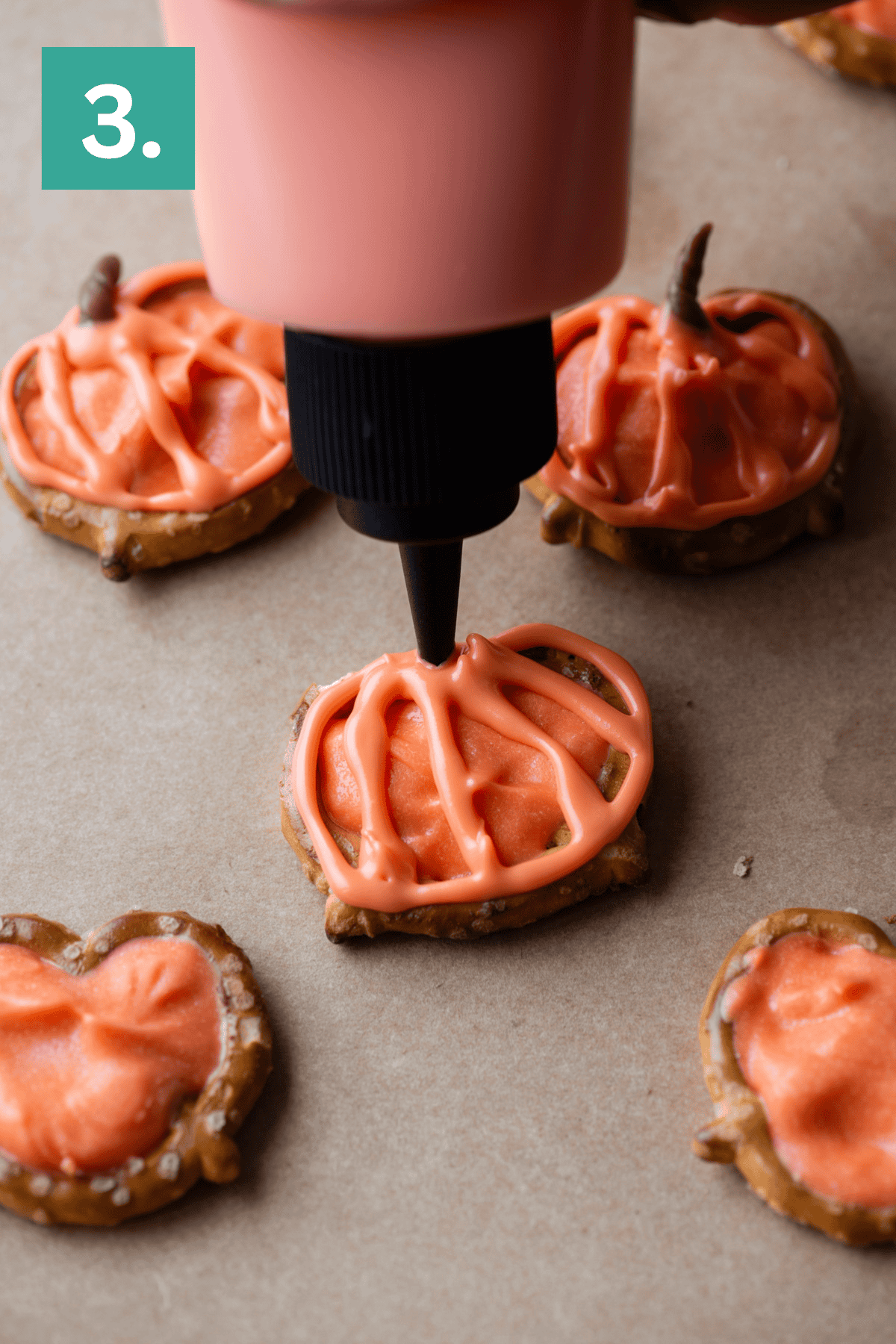 A hand uses a squeeze bottle to pipe orange icing onto a pumpkin-shaped cookie, with other iced cookies nearby on brown parchment paper. A green square with the number 3 appears in the top left corner.