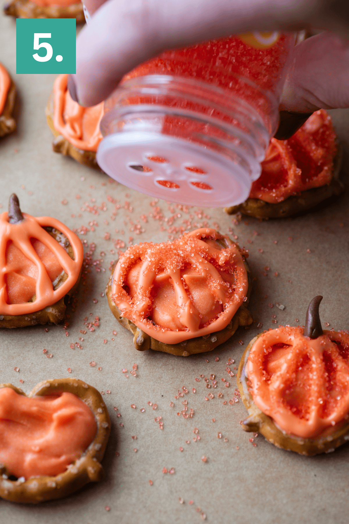 A hand sprinkles orange sugar over pretzel snacks decorated with orange icing to look like pumpkins on a sheet of parchment paper. A green box with the number 5 is in the upper left corner.