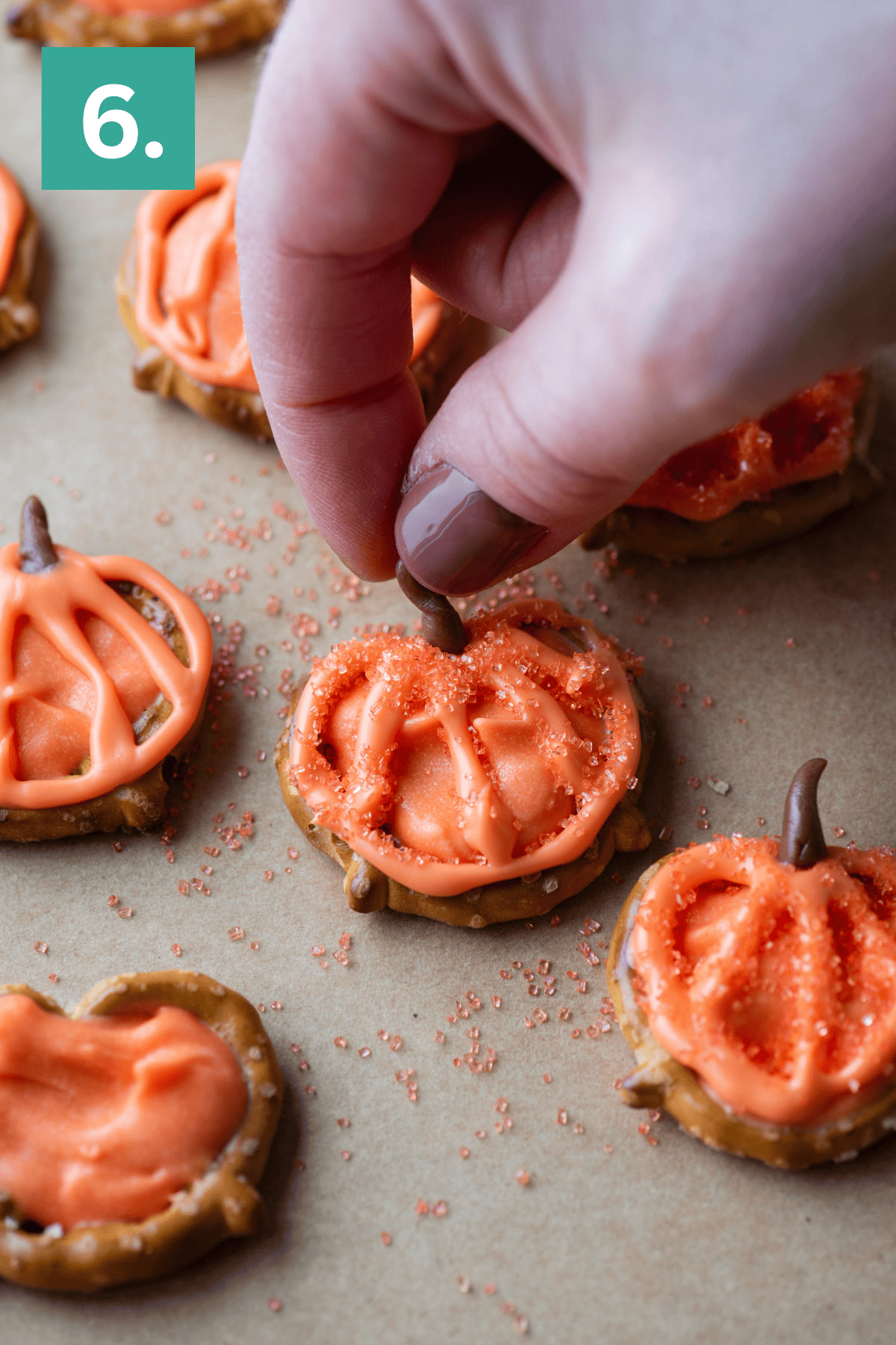 A hand places a small chocolate piece as a stem on a pretzel decorated with orange frosting to look like a pumpkin. Other similar pumpkin-shaped pretzel treats are on a sheet of parchment paper.